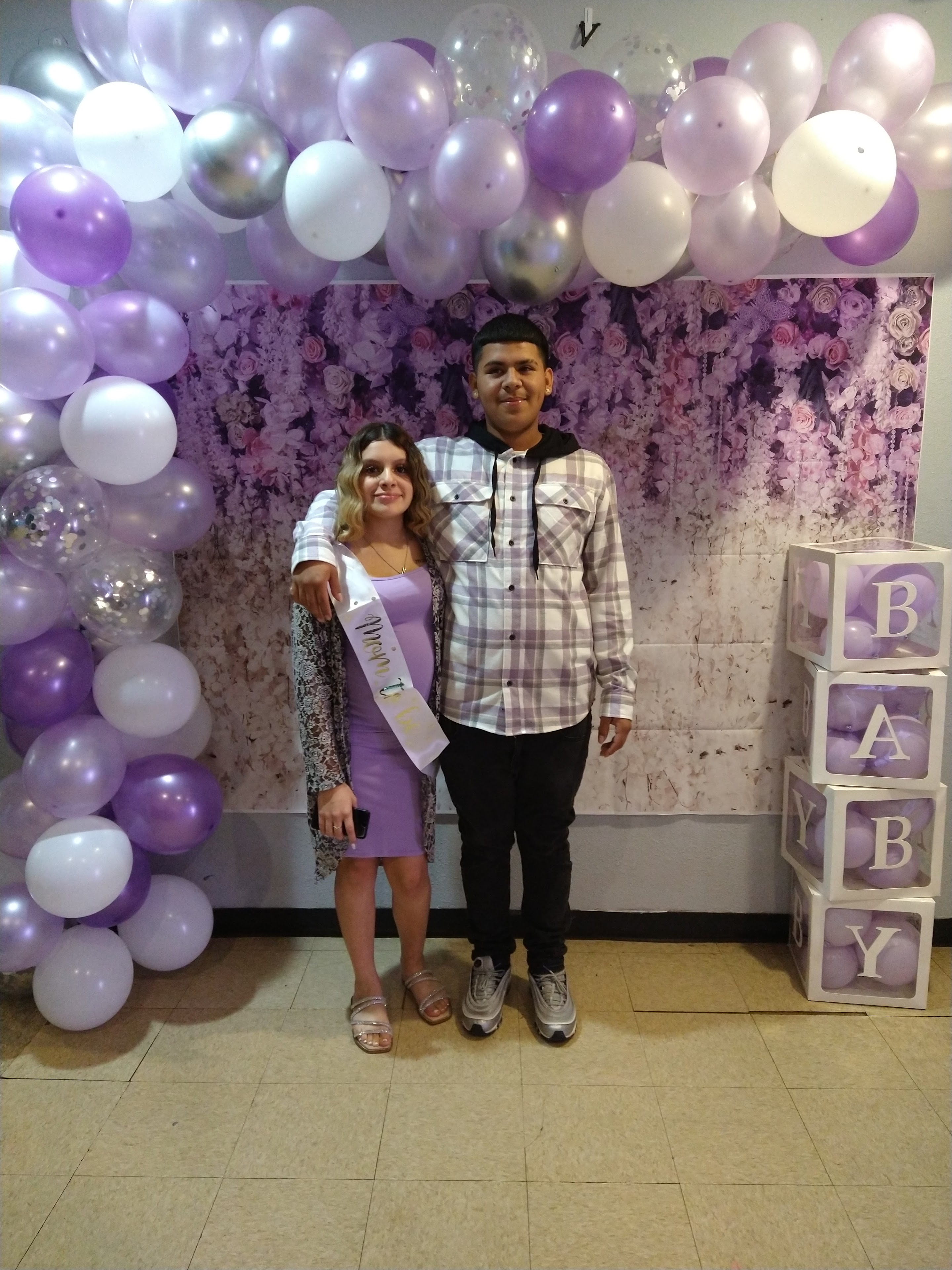 A couple joyfully poses at a baby shower with purple balloons celebrating a new arrival.