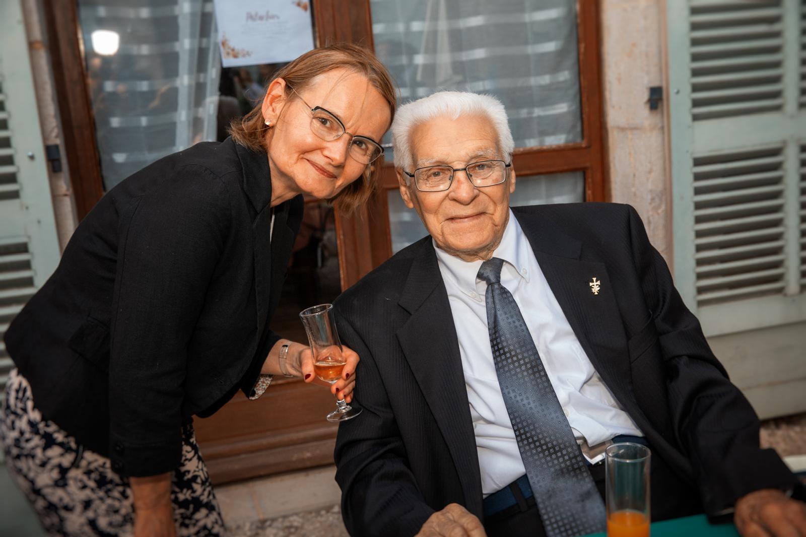 A gentleman in formal attire poses with a woman at a gathering, enjoying drinks outdoors.