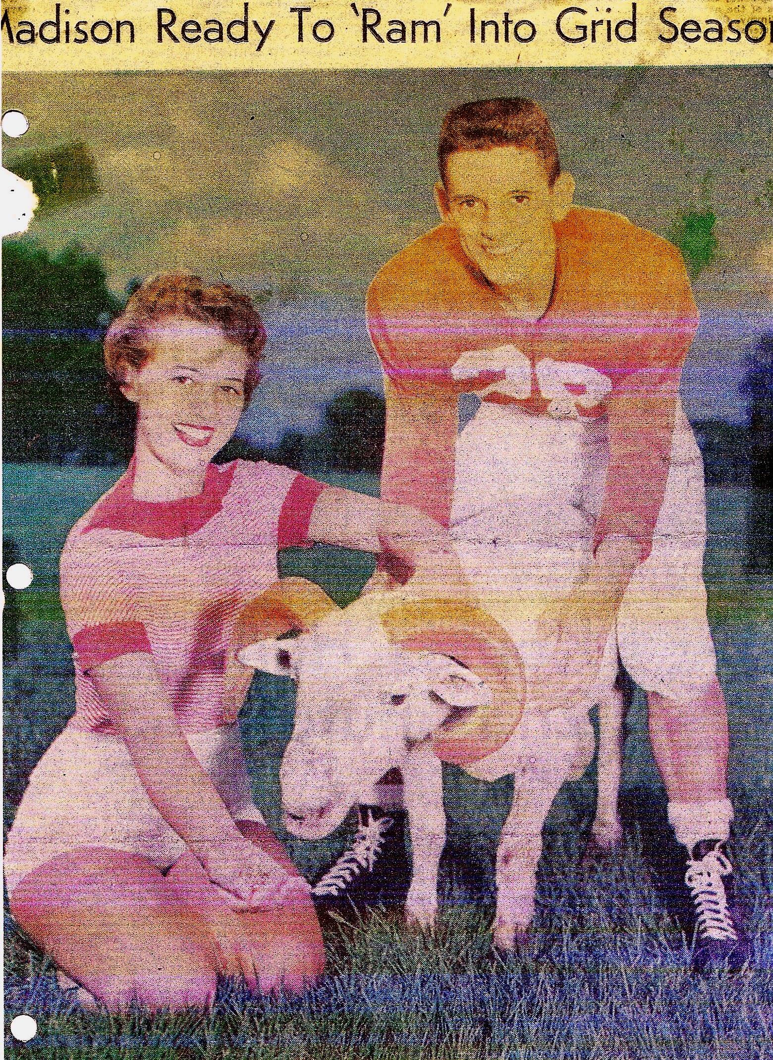 A cheerful couple interacts with a ram in a sunny field, showcasing a joyful moment together.