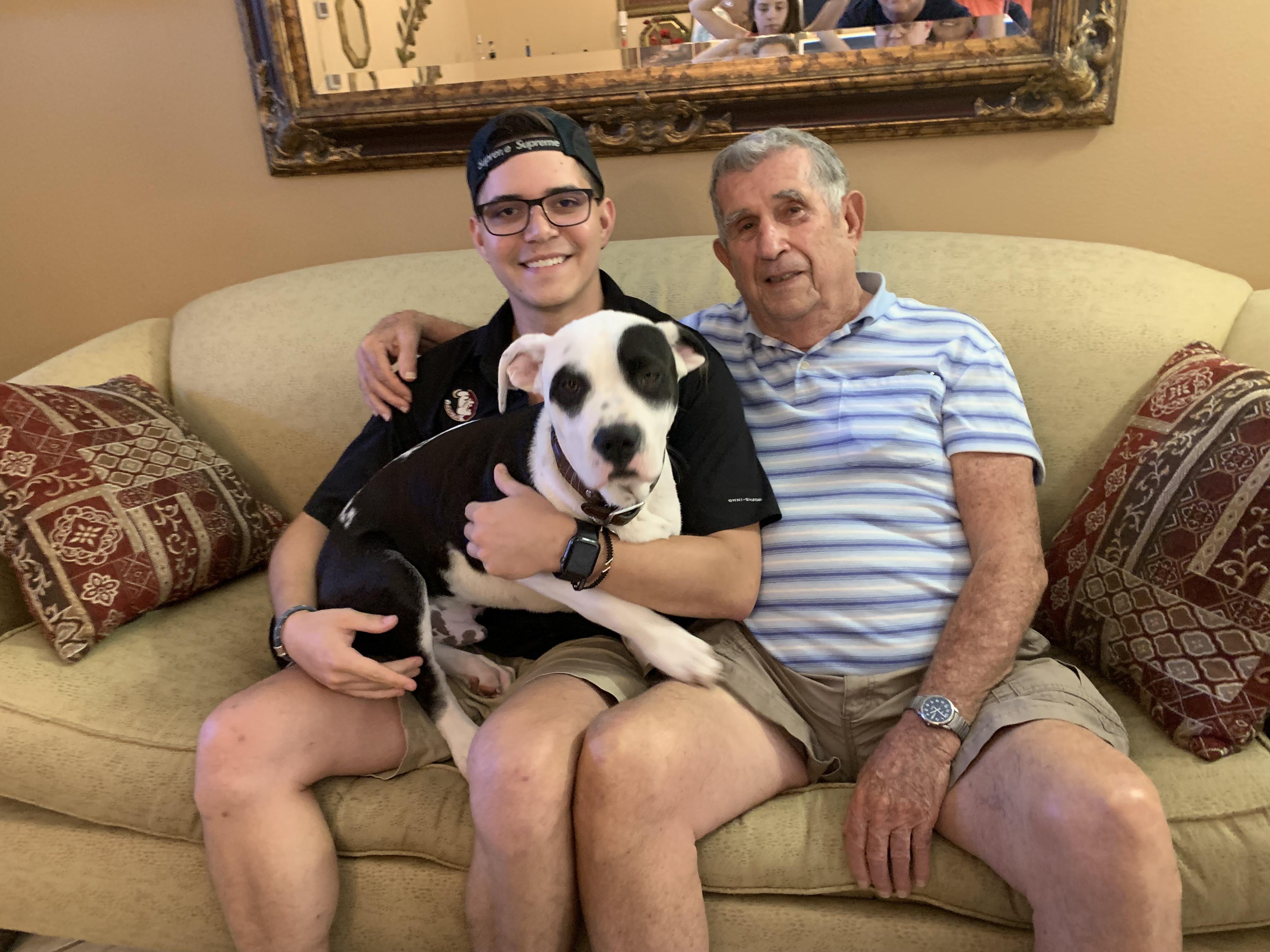 Two men sit on a couch with a dog, enjoying a relaxed moment together in a warm living room.
