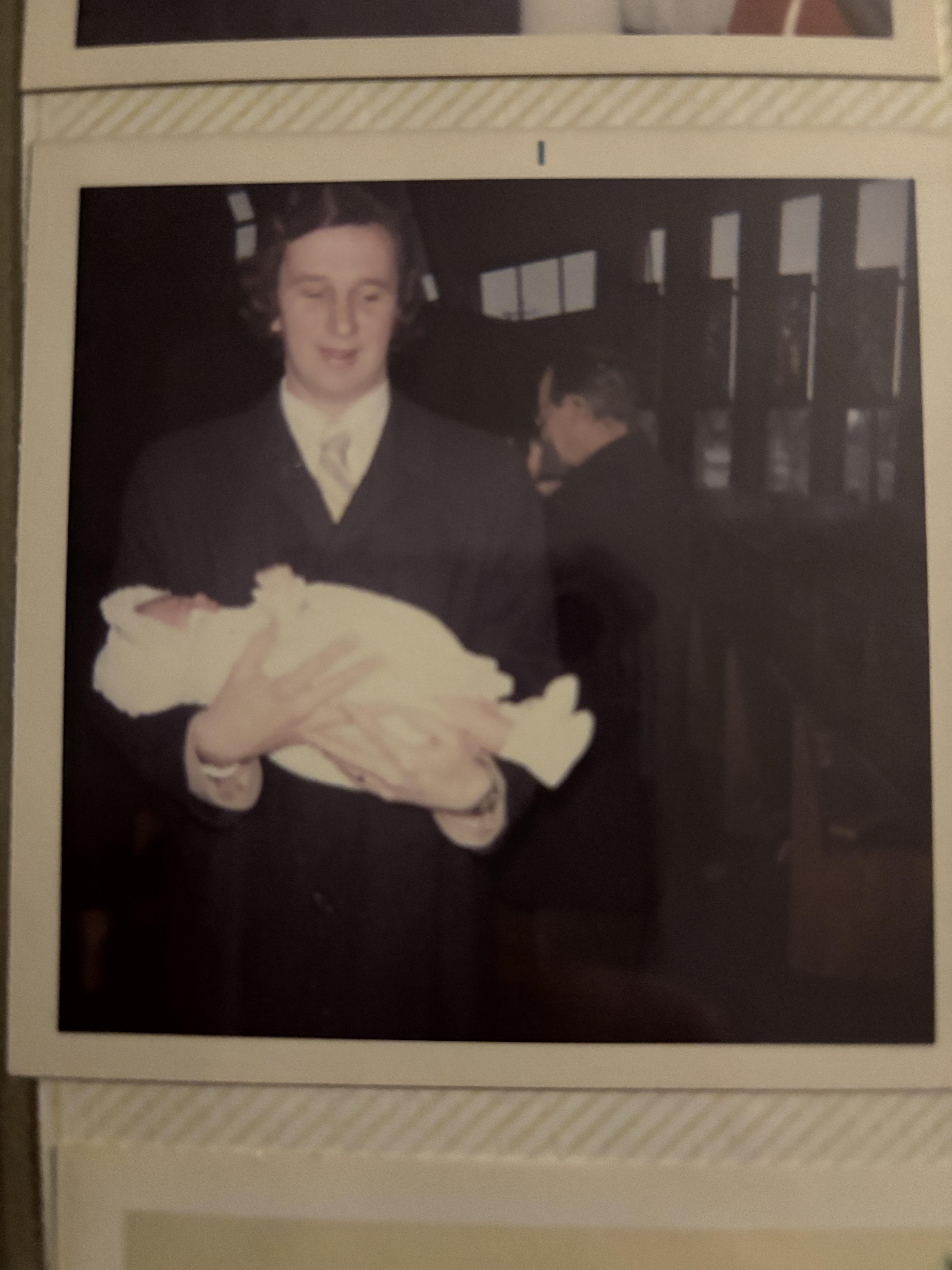 A woman stands holding a newborn baby, dressed in formal attire, in a dimly lit interior.