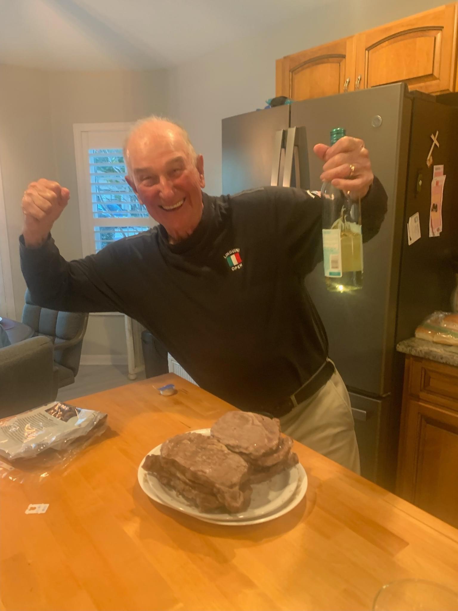 A joyful chef poses with wine and steak in a welcoming kitchen during a casual dinner gathering.
