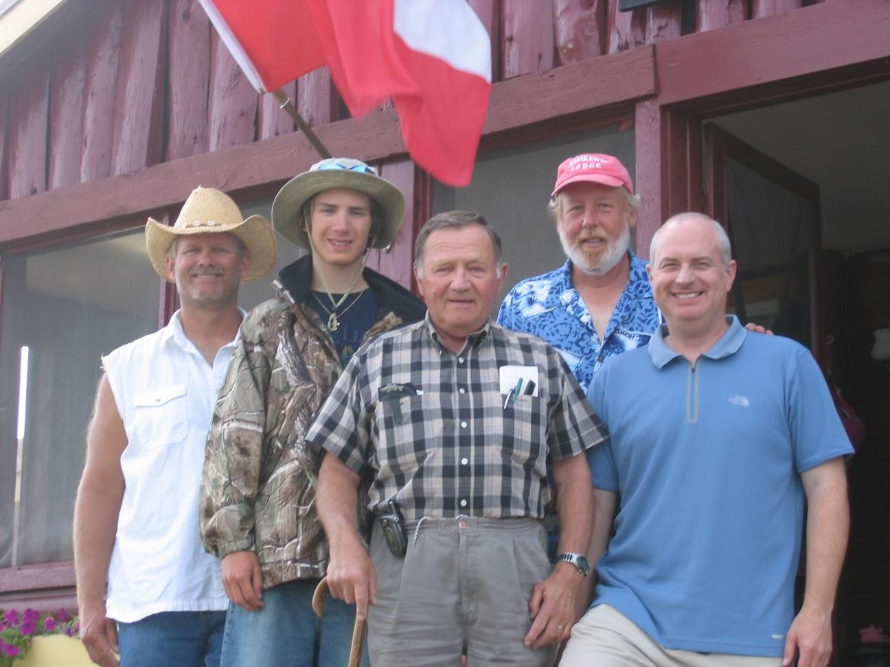 Men gather outside a building, proudly posing with a flag on a sunny summer day in a rural area.