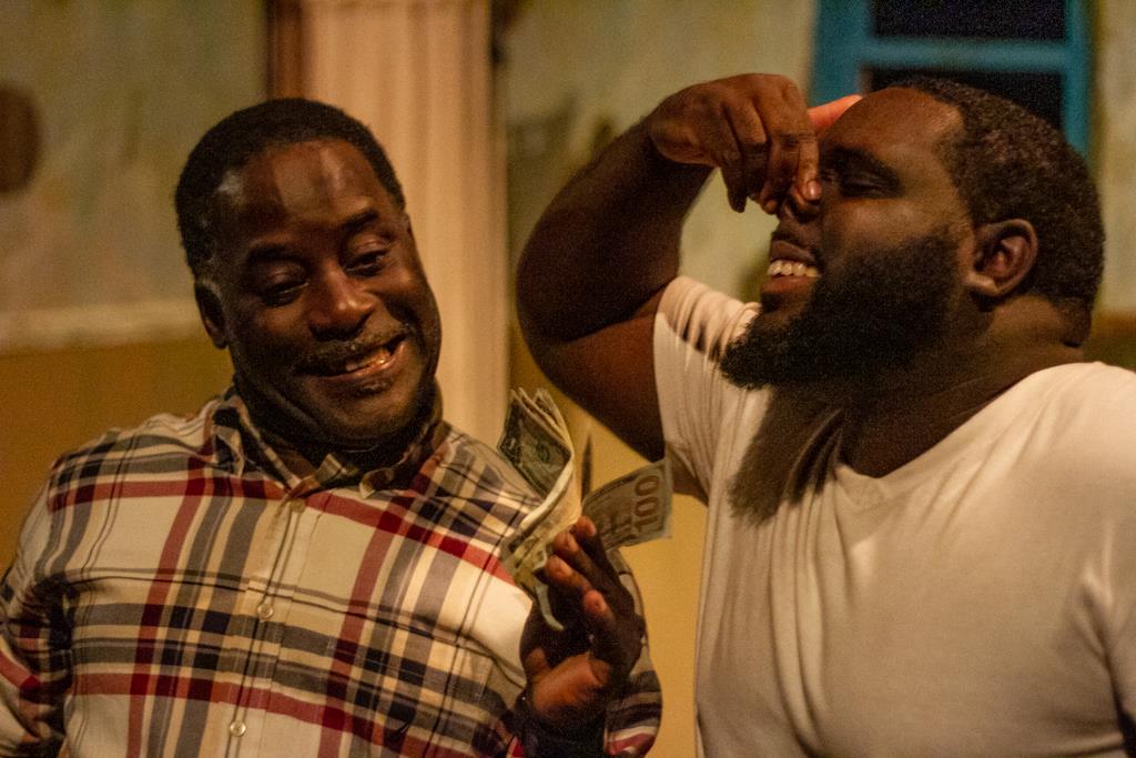 Two friends express joy with laughter while one playfully holds cash in a cozy room at night.