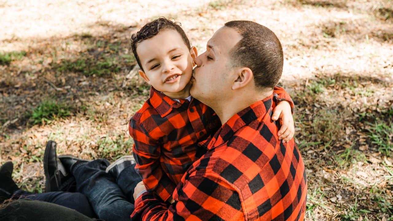 A father lovingly kisses his son while they sit on the grass, both wearing matching outfits.