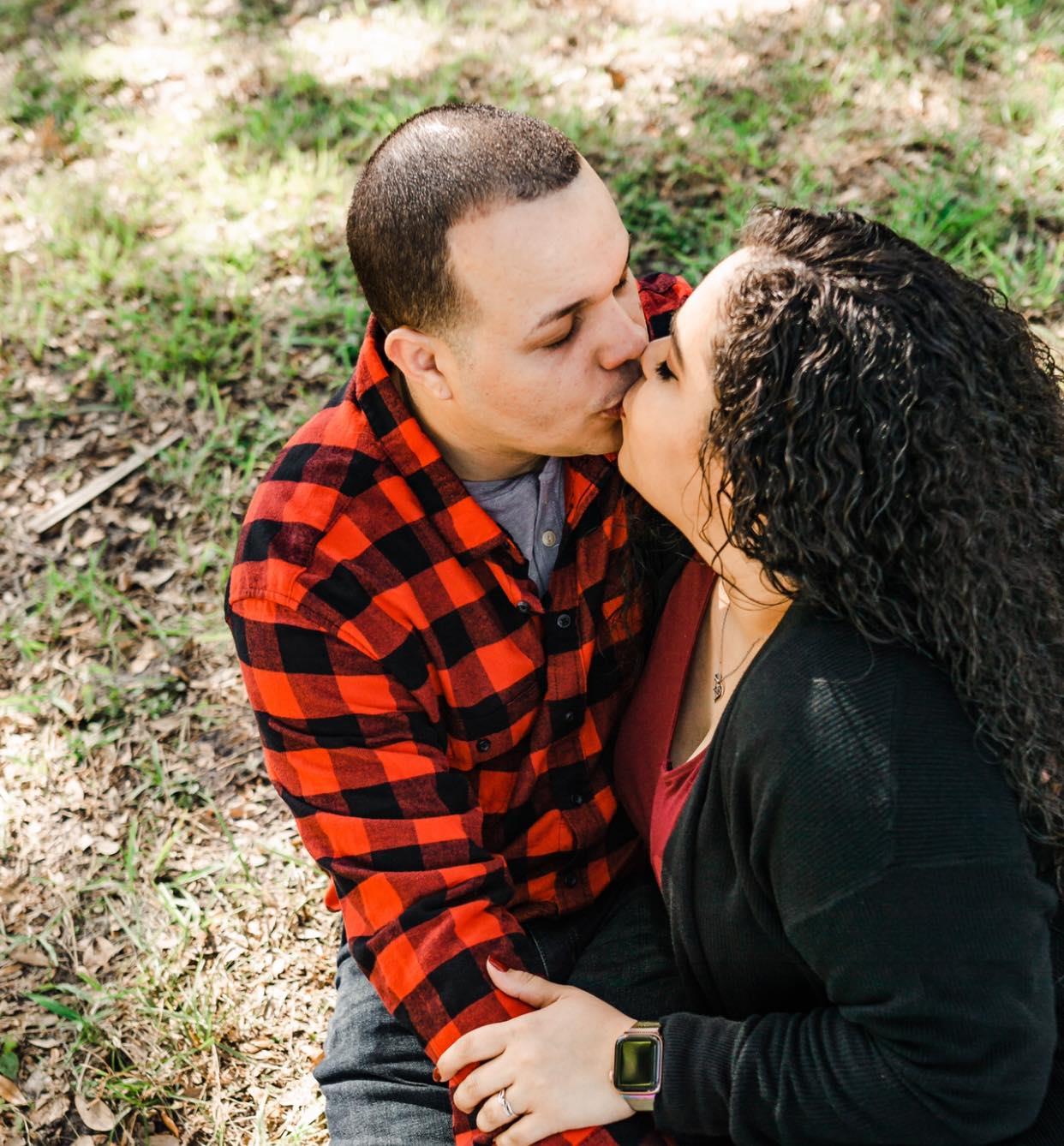 Two people embrace affectionately, sharing a kiss in a lush green park under sunlight.