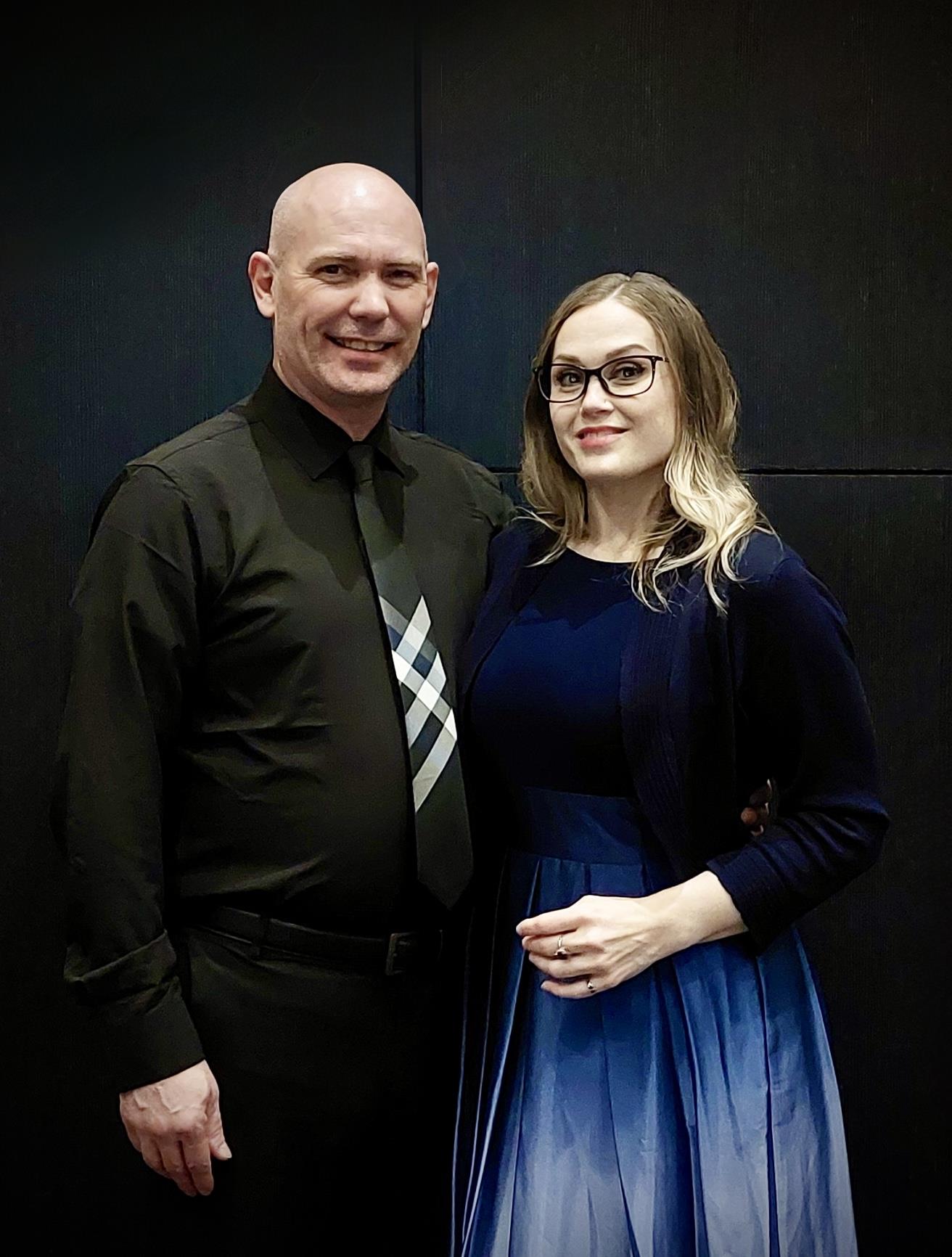 A couple dressed in formal attire standing together, smiling with a dark background.