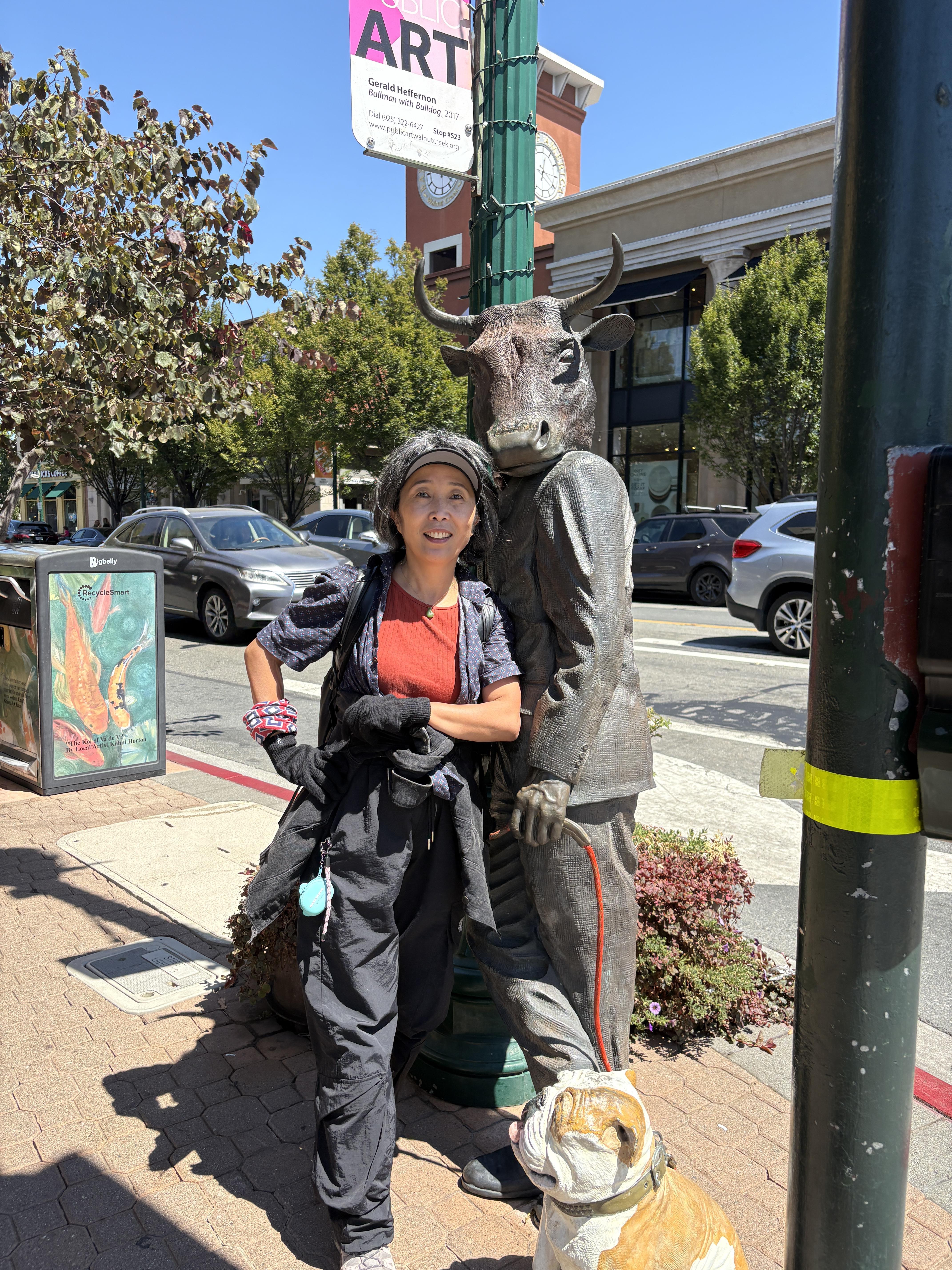 A woman happily stands next to a cow statue on a vibrant urban street filled with cars.