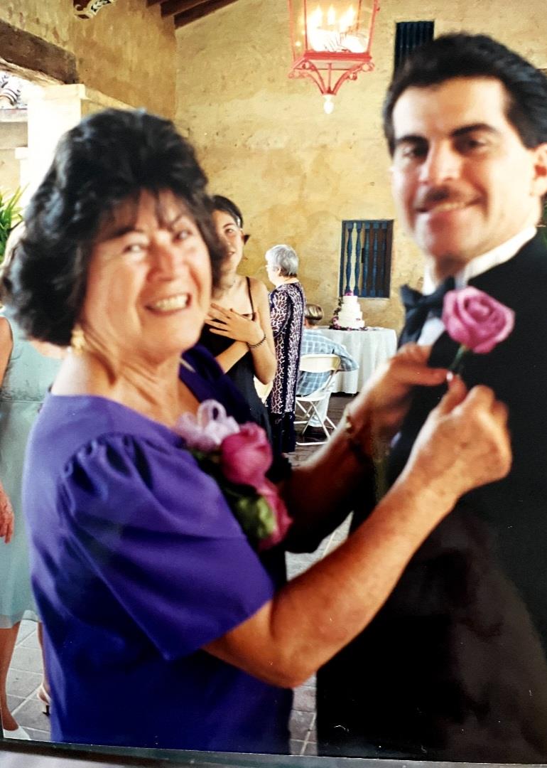 A woman adjusts a boutonniere on a man while smiling, surrounded by guests at a wedding.