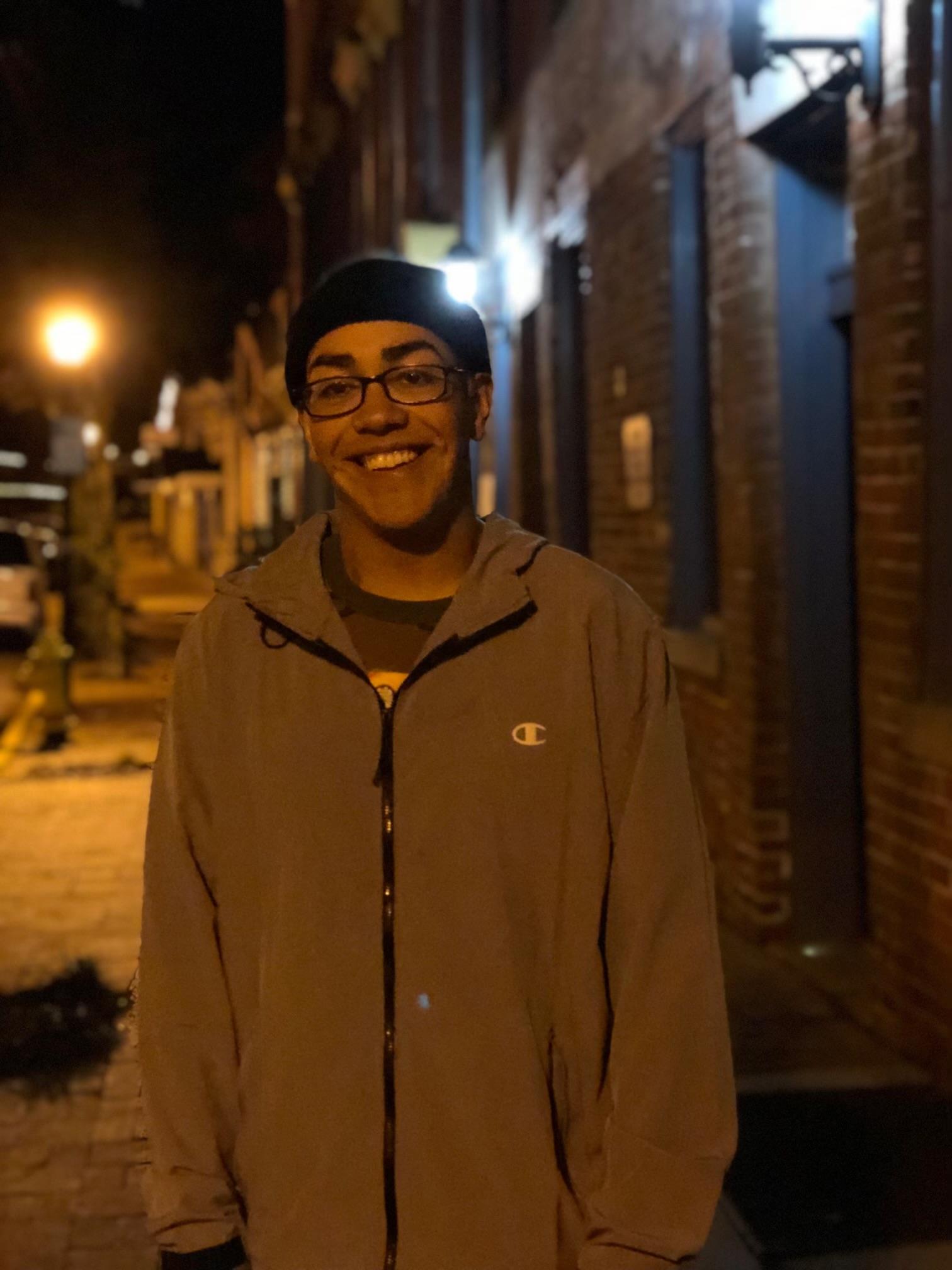 A young man stands against a brick wall, smiling brightly on a dimly lit street at night.
