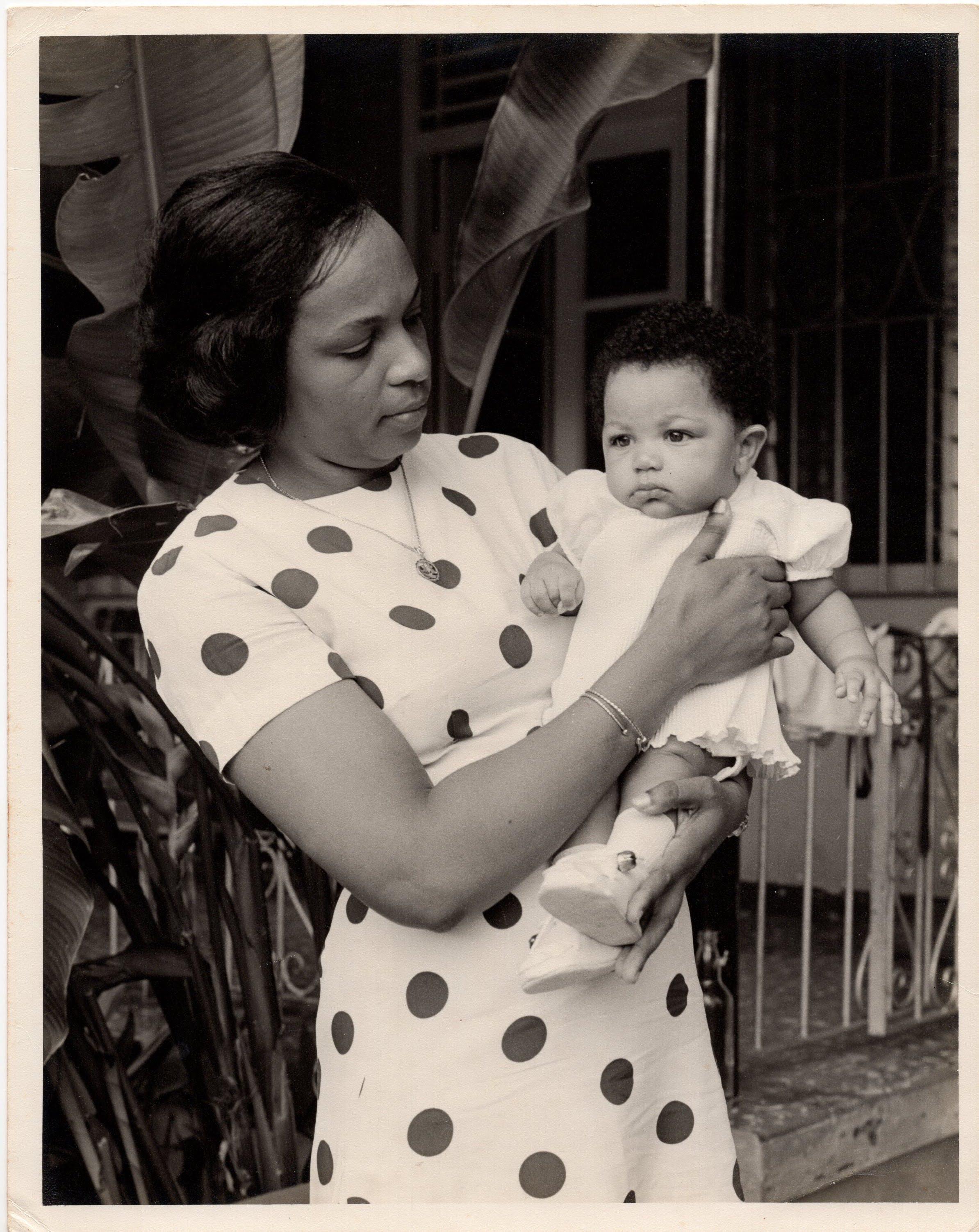 A woman lovingly cradles her baby in a garden, enjoying a bright day with greenery around them.
