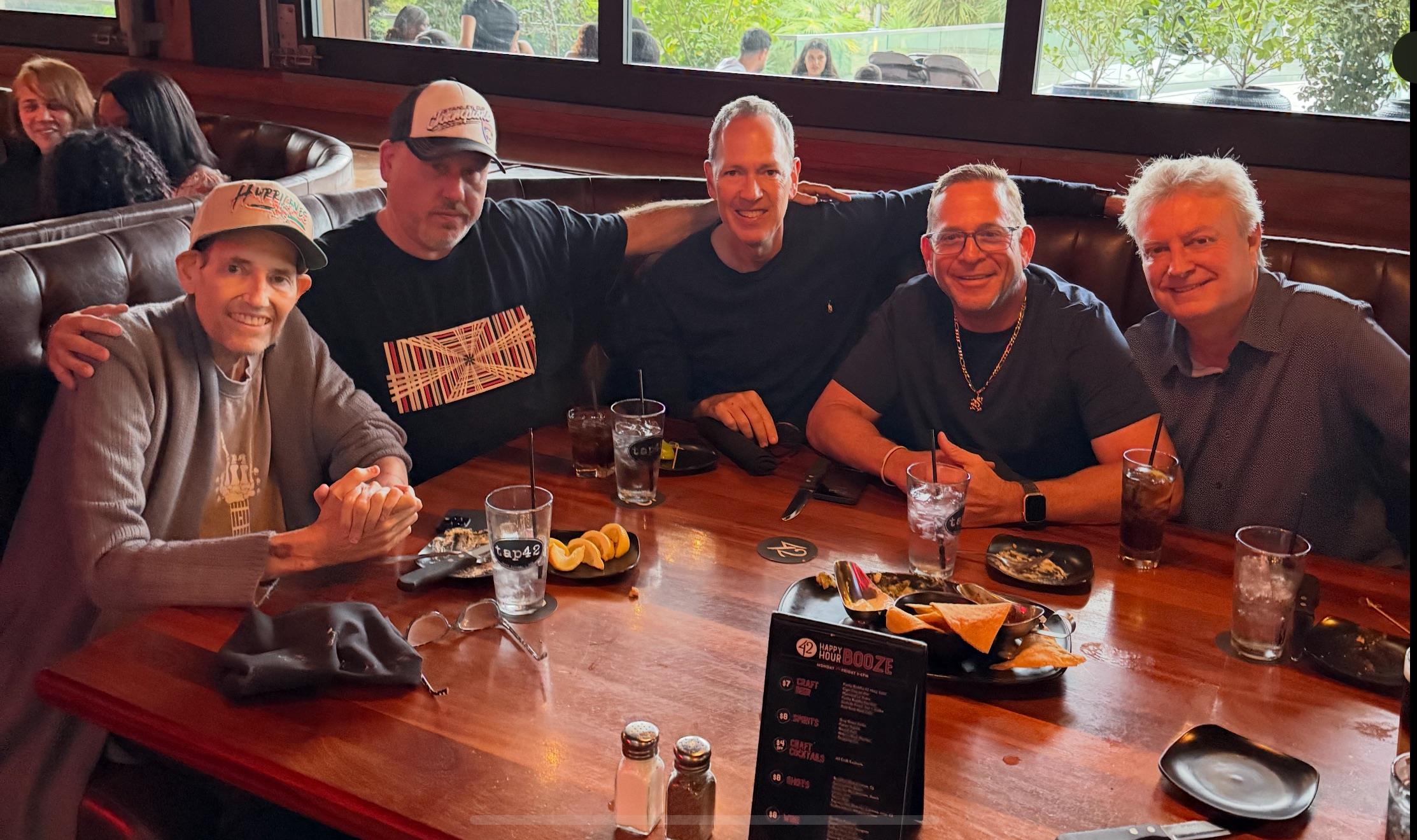 Four friends sit around a table, enjoying food and drinks while smiling and chatting.
