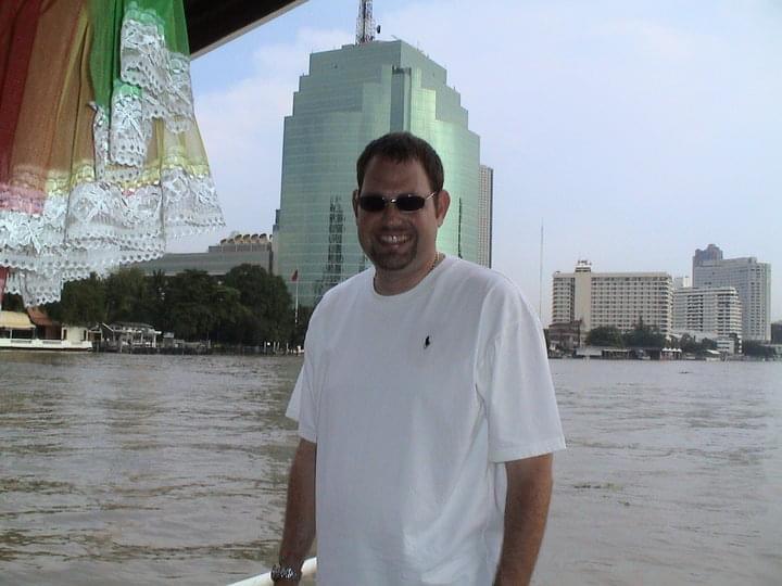 A man stands on a boat enjoying a sunny day by the river with a city skyline in the background.