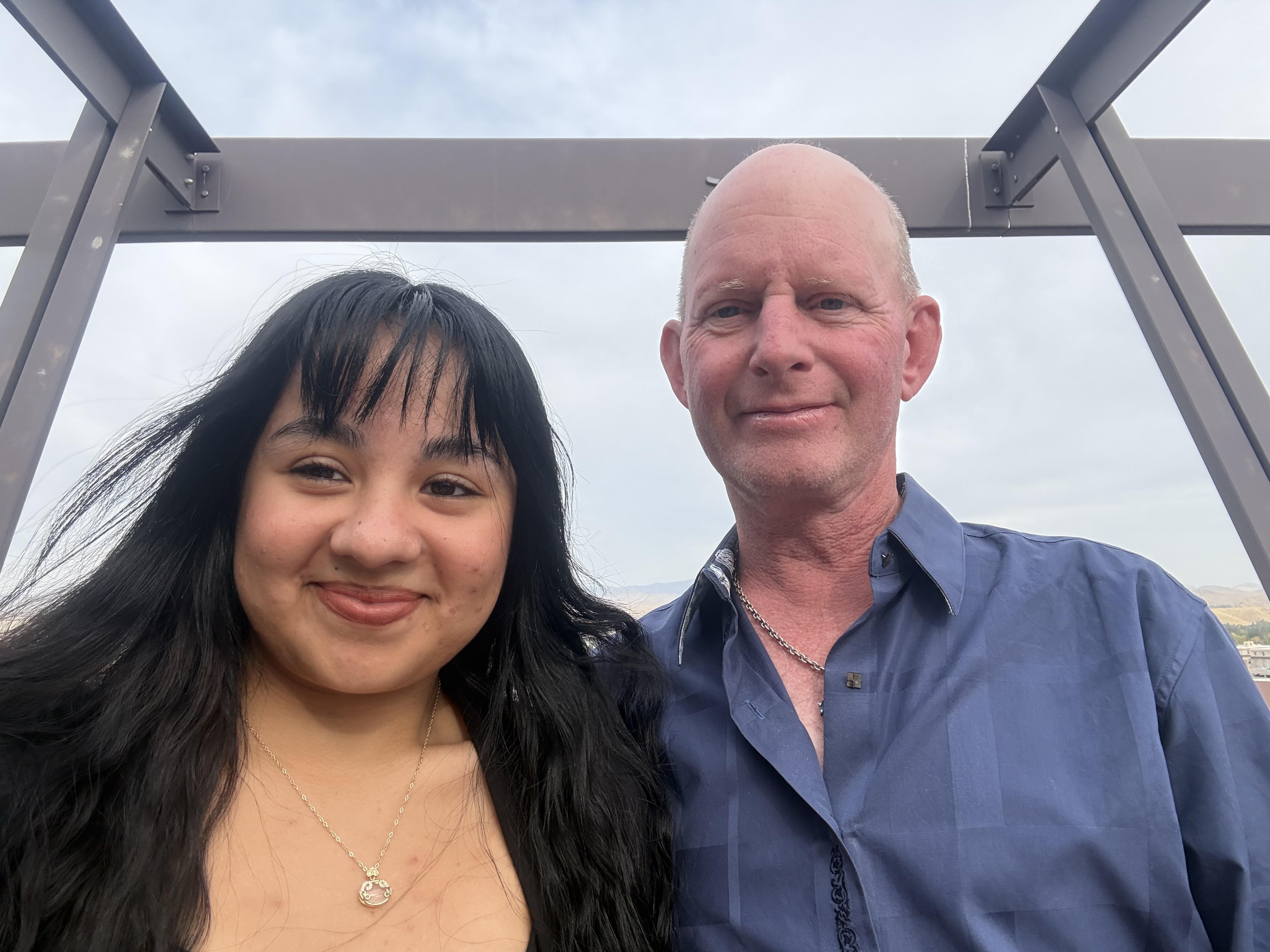 Two people are joyfully posing outdoors at a contemporary structure under a clear sky.