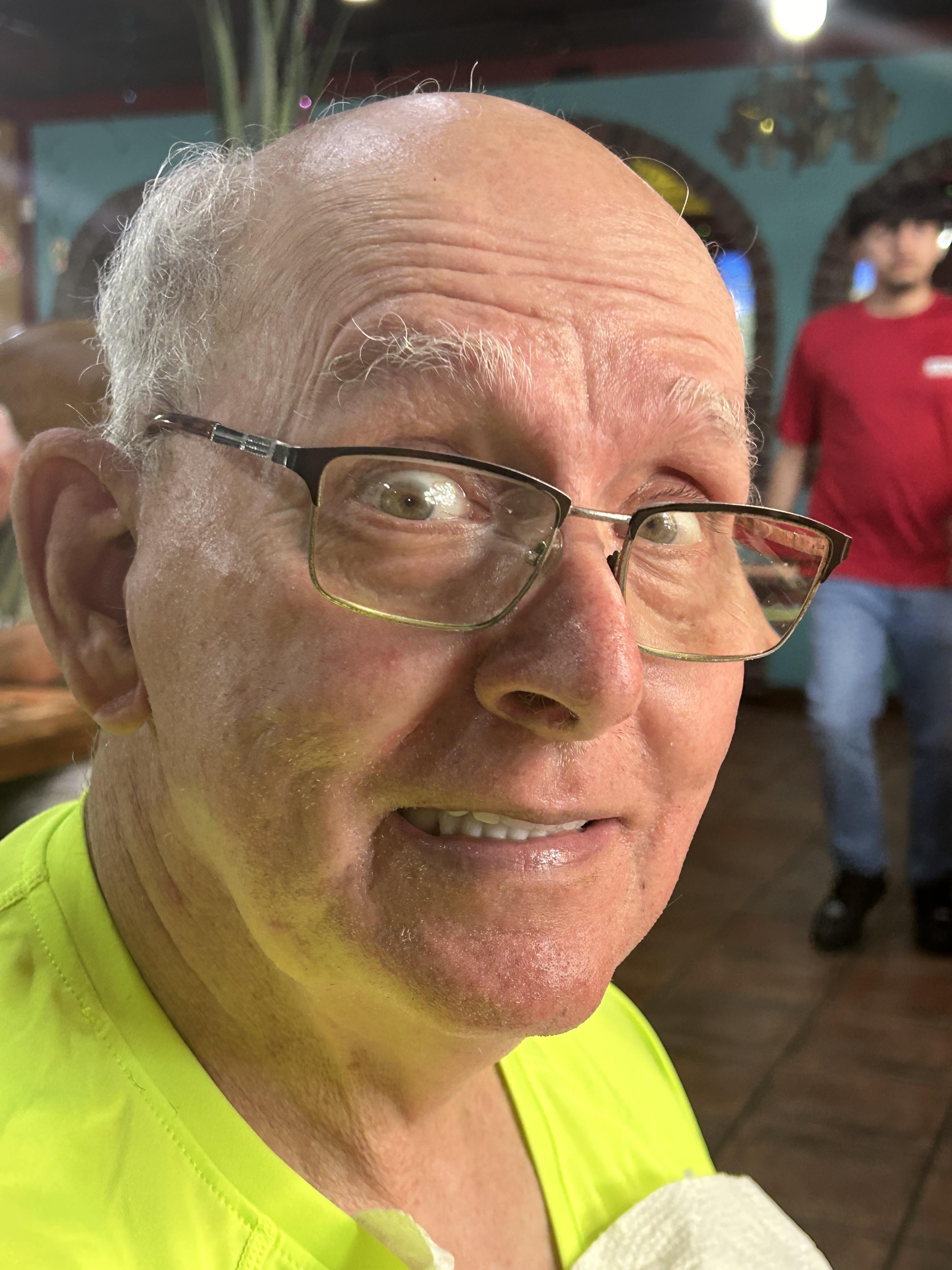 An elderly man in bright clothing smiles while interacting with others in a bustling space.