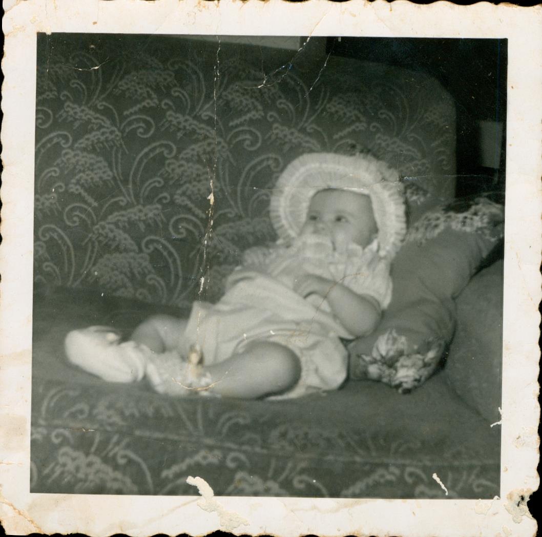 A baby sits comfortably on an old couch, wearing a soft hat and holding a toy, seemingly at ease.