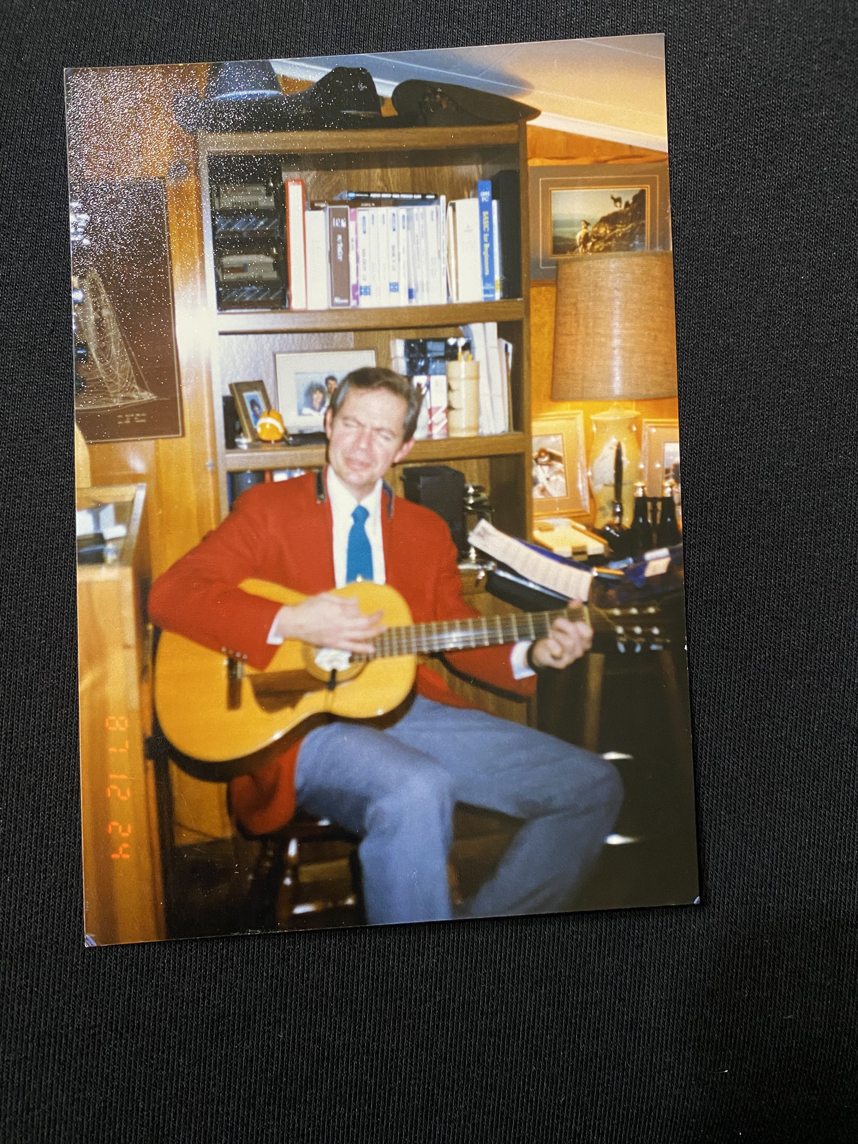 A man sits in a cozy office, strumming a guitar and smiling, dressed in a red jacket and blue tie.