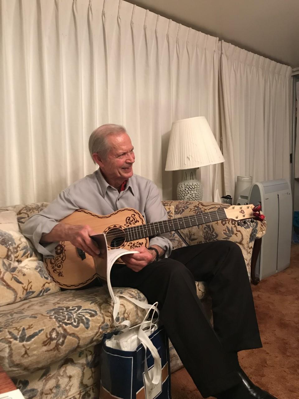 A joyful elderly man strums a guitar while sitting comfortably in a warm, inviting living room.