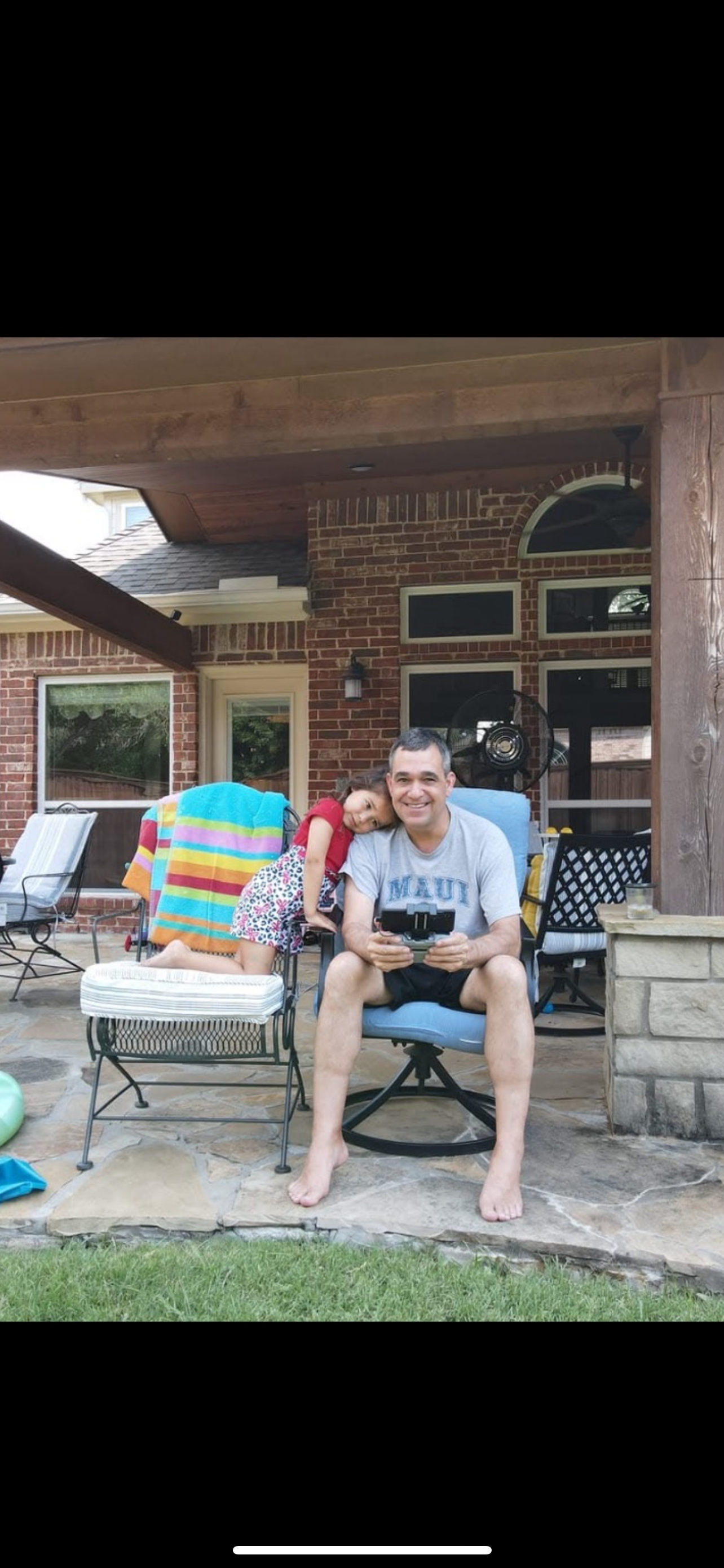A child leans on an adult while sitting outside in a backyard on a sunny day.