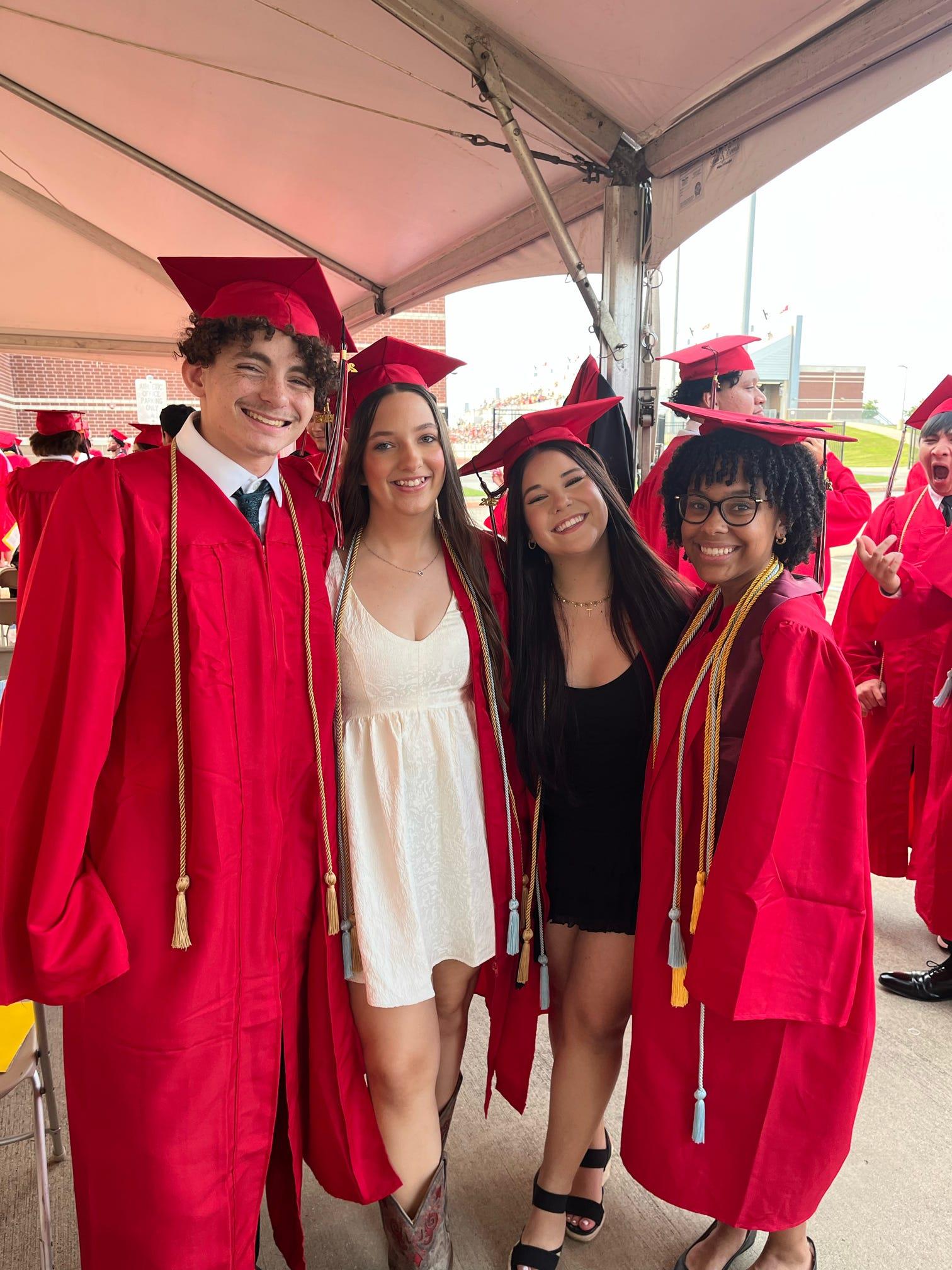 Four graduates in red gowns smile together at their graduation ceremony in the sun.