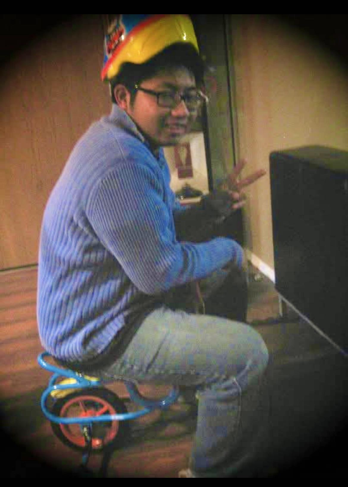 Individual wearing glasses and a sweater enjoys sitting on a tricycle indoors, smiling and posing.