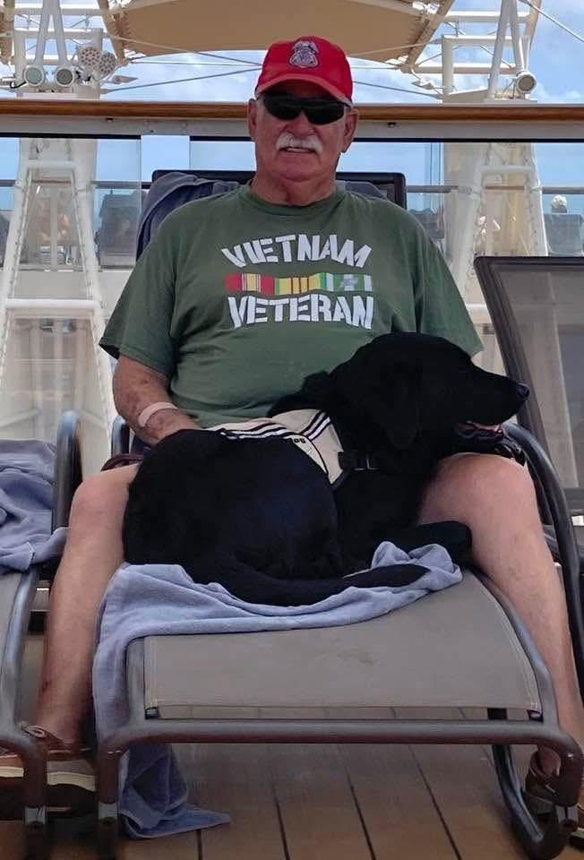 An elderly man in a green shirt sits on a lounge chair with a black dog beside him.