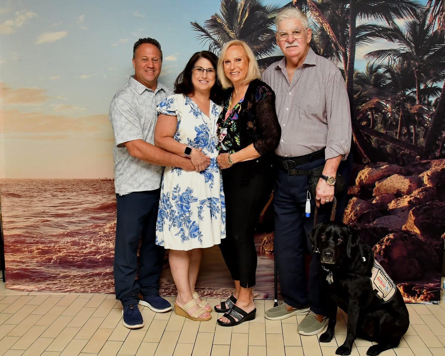 Four individuals and a dog pose together against a tropical backdrop at a summer event.