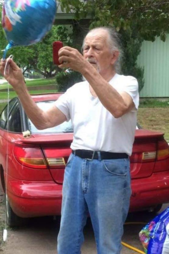 An elderly man in a white shirt and blue jeans stands by a red car, happily taking a selfie.
