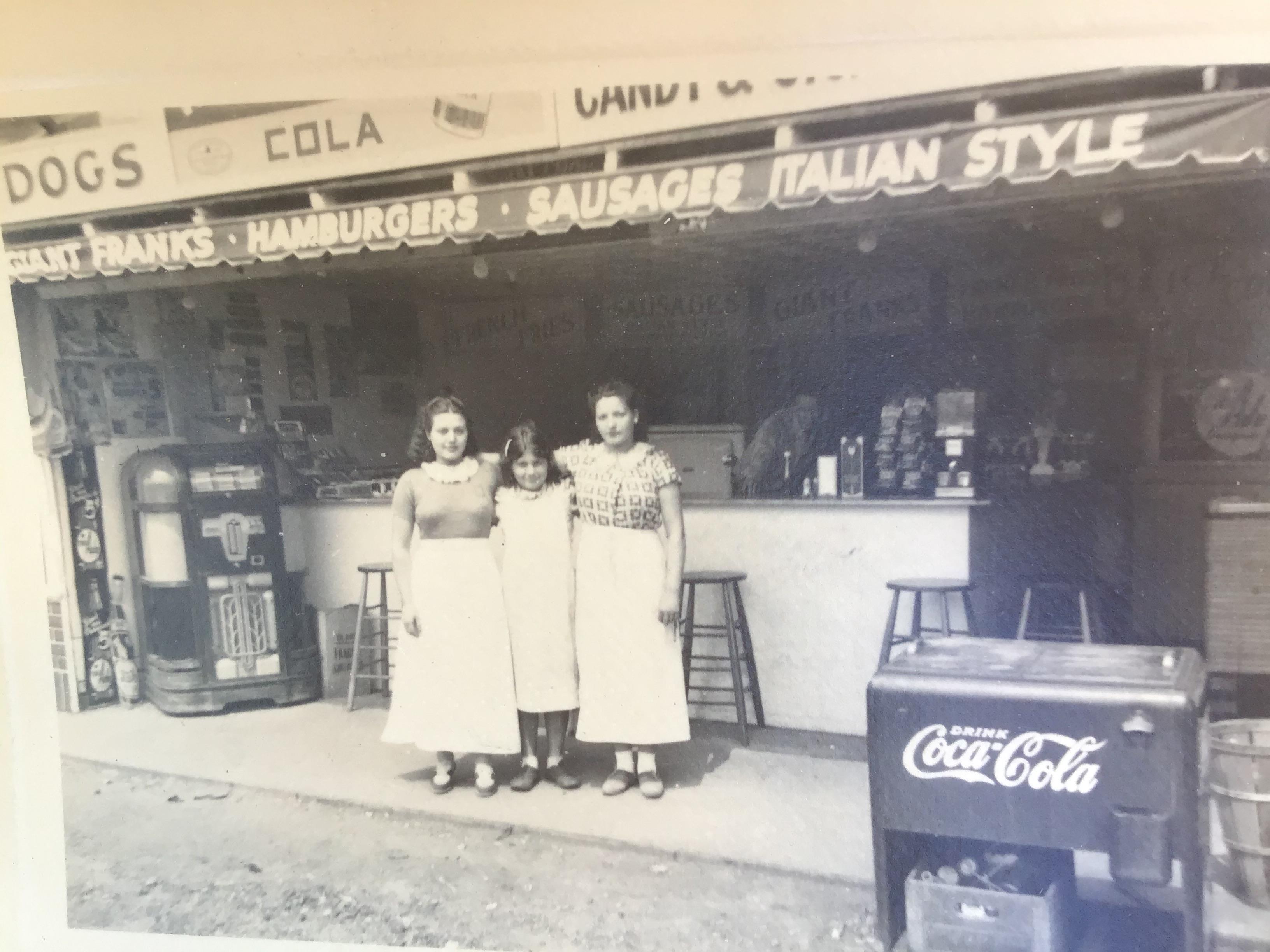 Three women in period uniforms pose in front of a quaint diner with nostalgic signage.