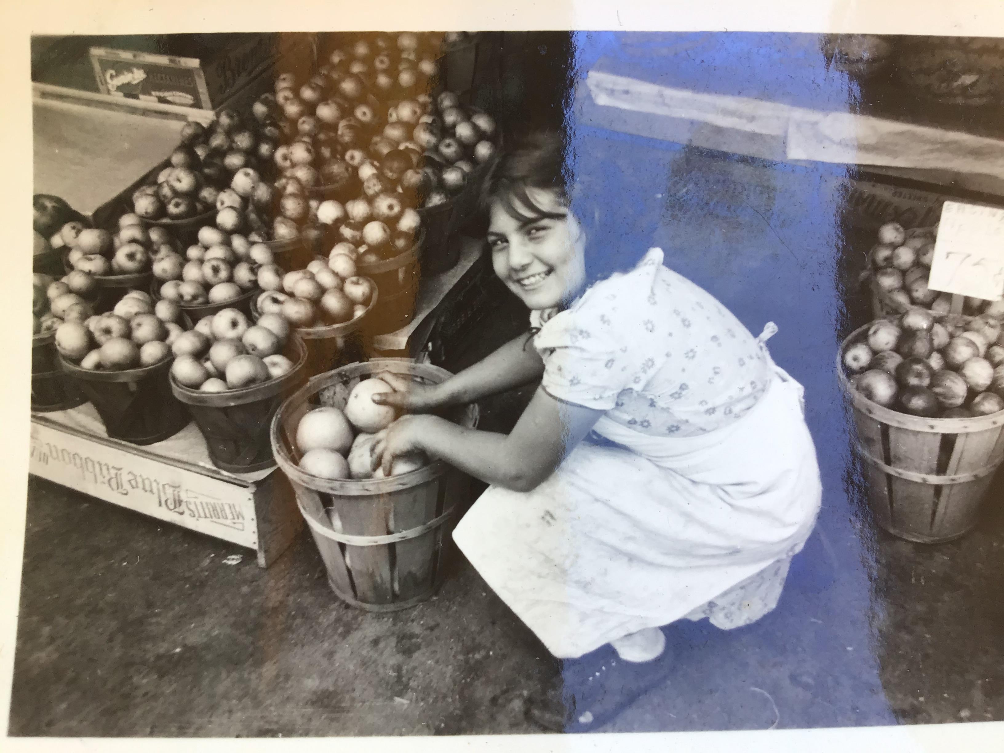 A girl happily collects apples from a basket in a vibrant market setting filled with fruit.