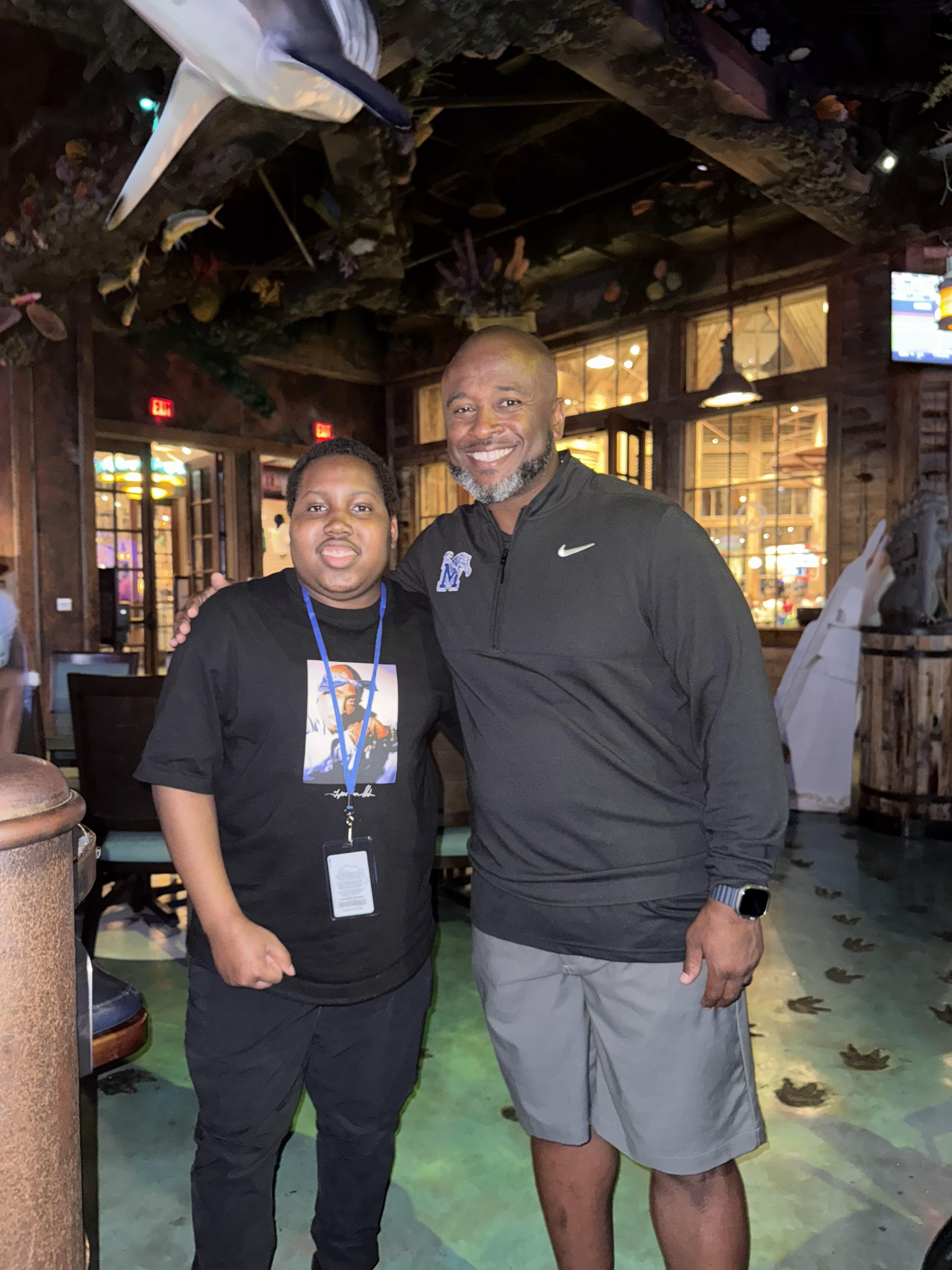 Two men stand smiling inside a lively restaurant decorated with natural elements.