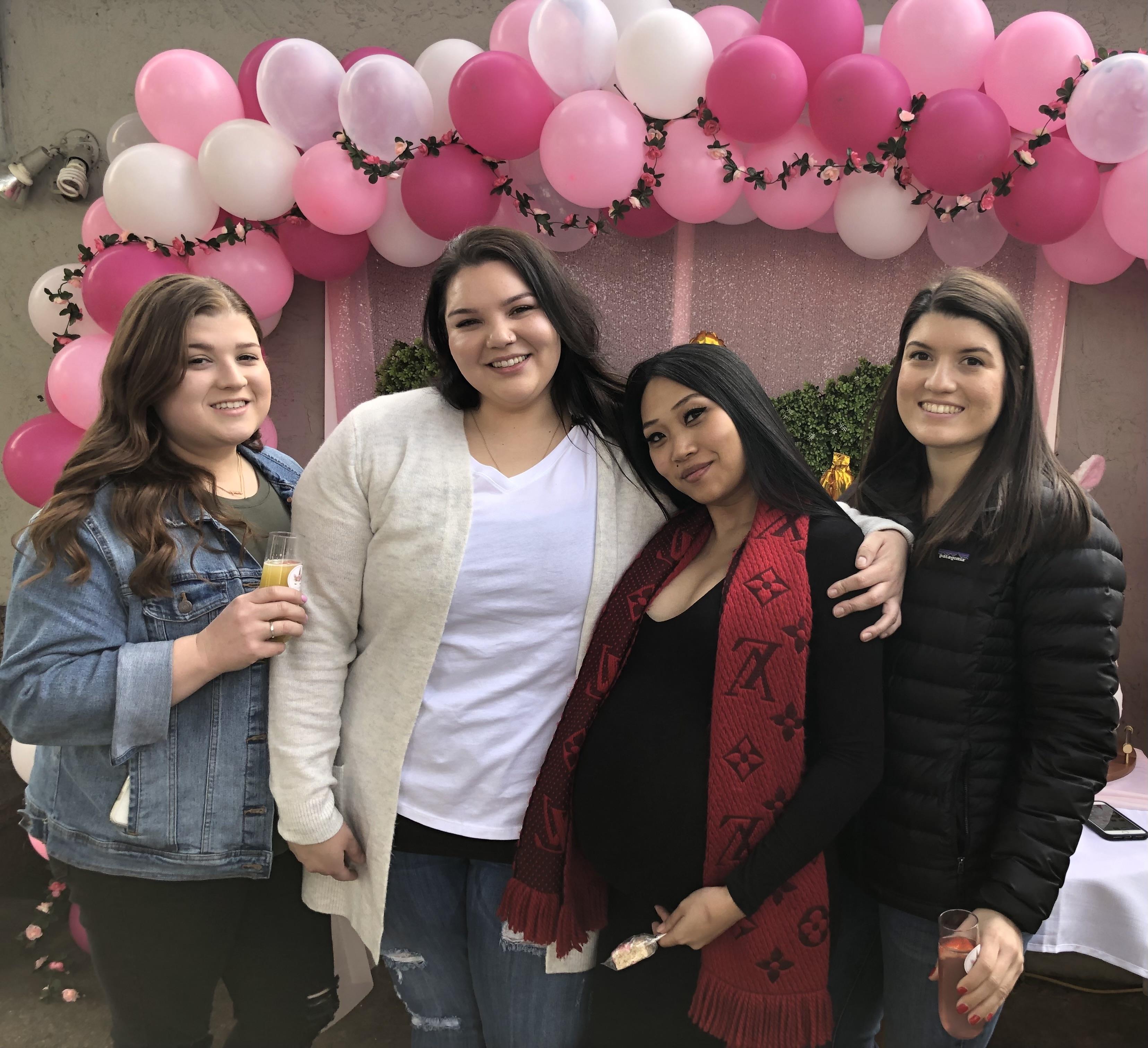 Four friends smile near pink balloons, enjoying a cheerful moment at a daytime event.