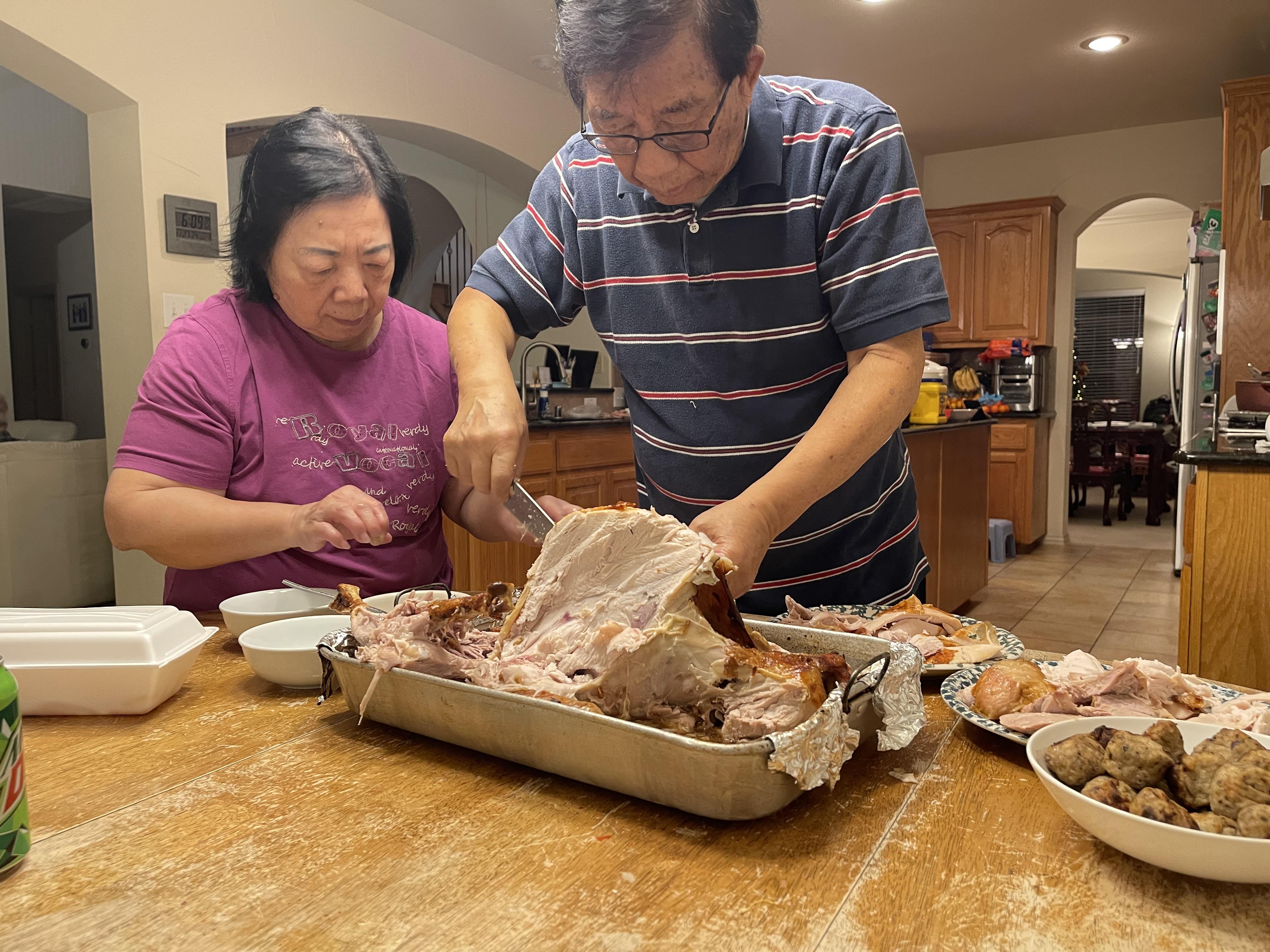 Two family members carve a turkey while preparing side dishes at a wooden dining table.