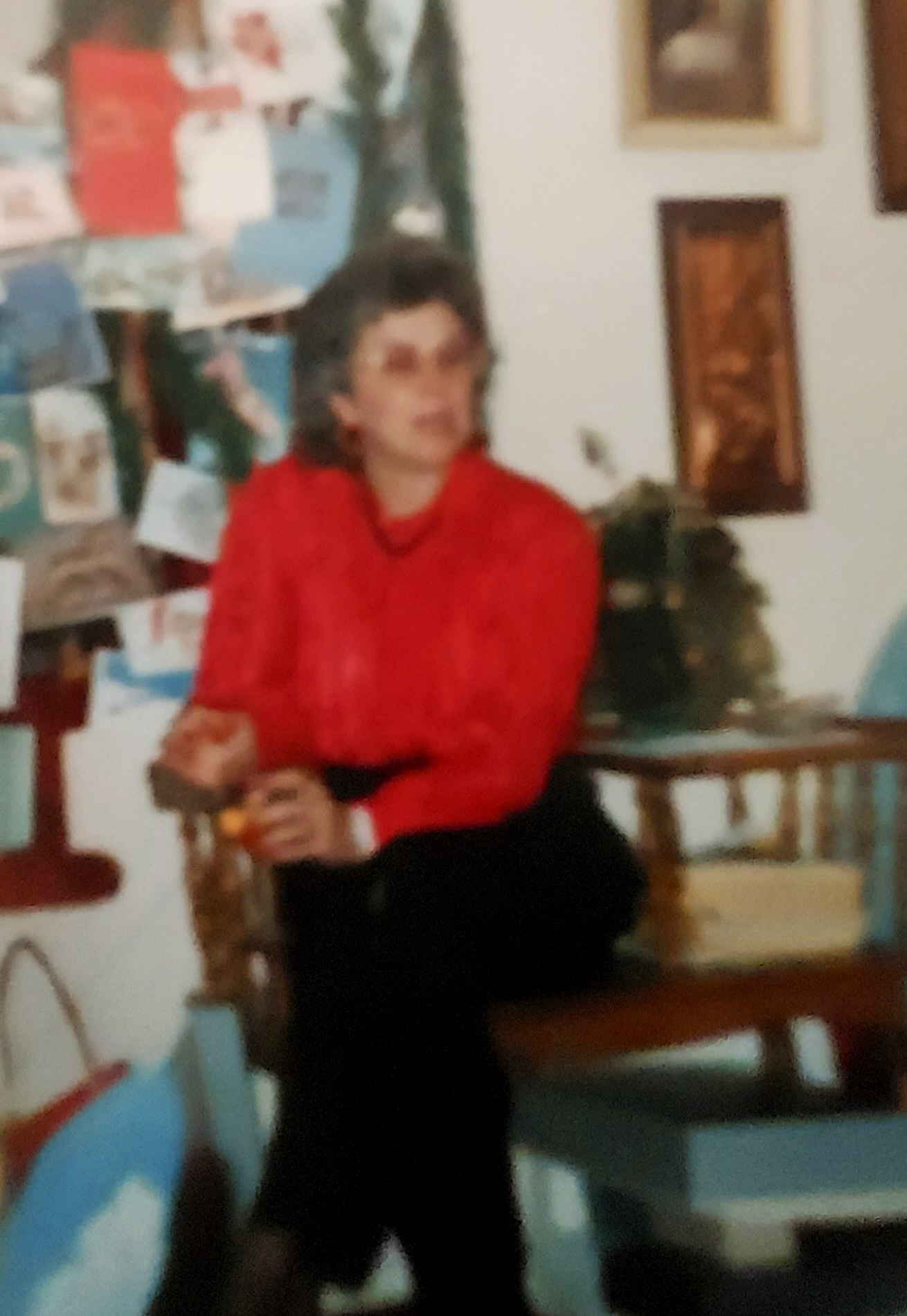 A woman wearing a red blouse is seated on a wooden bench, holding an object and looking thoughtful.