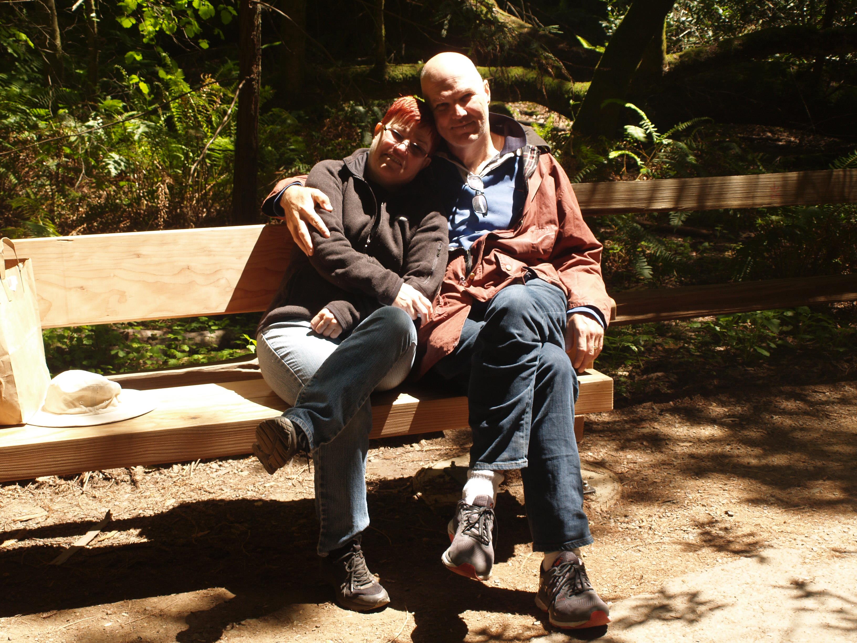 A couple sits closely on a bench, smiling and enjoying a warm day among lush greenery.