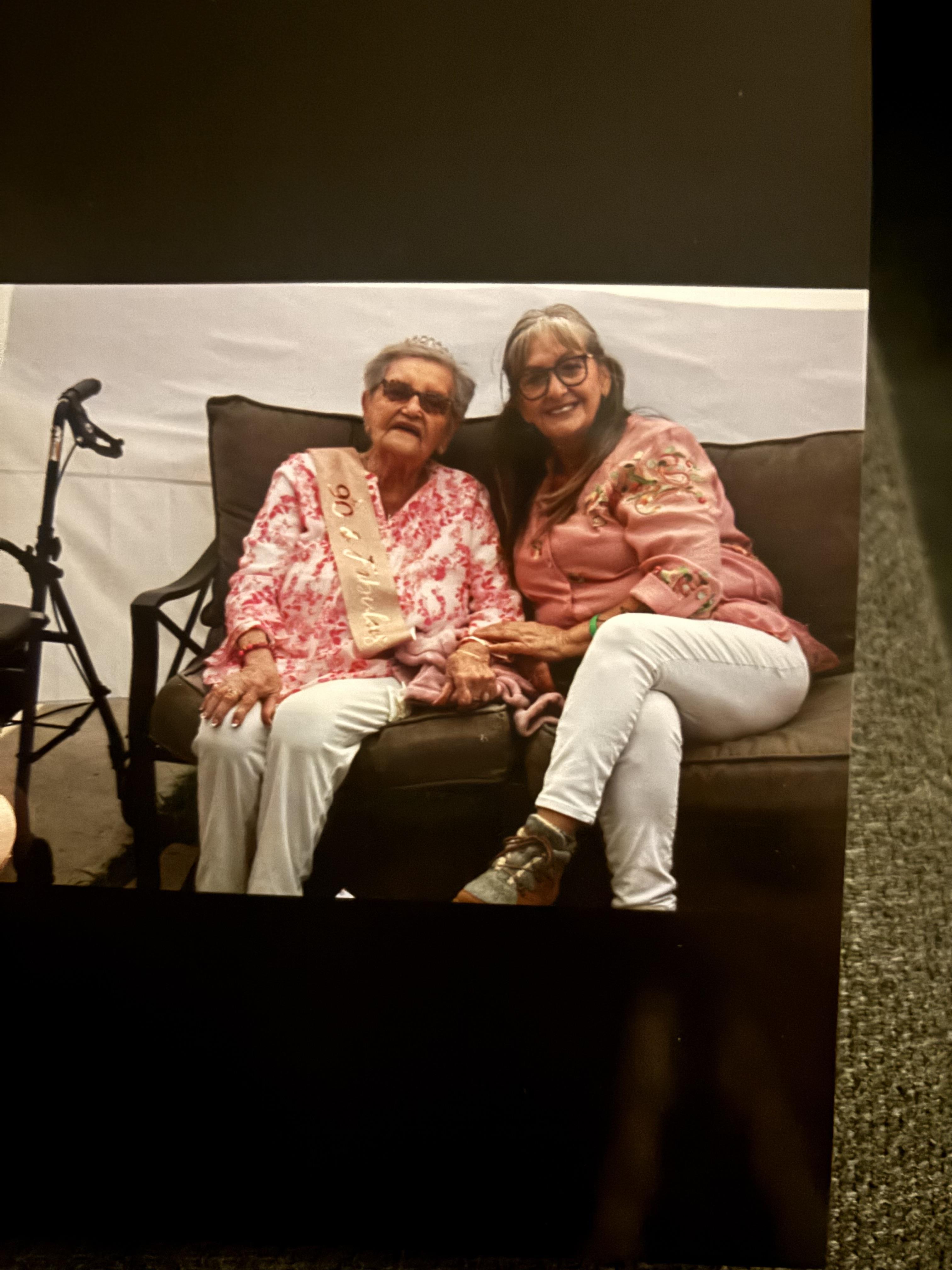 Two women sit closely on a sofa, smiling and enjoying each other's company in a cheerful setting.