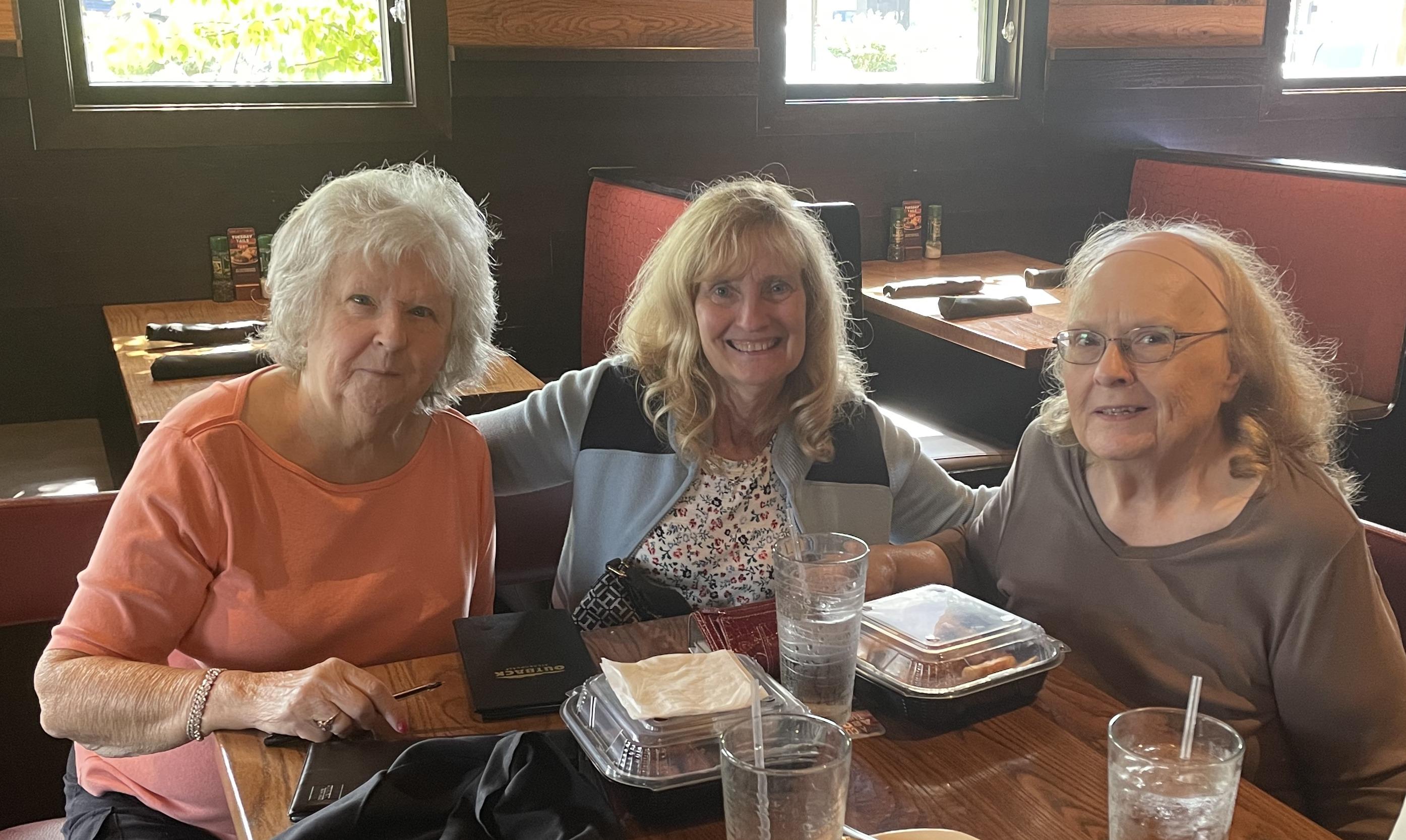Three friends smile happily while sitting at a table with drinks and takeout containers.