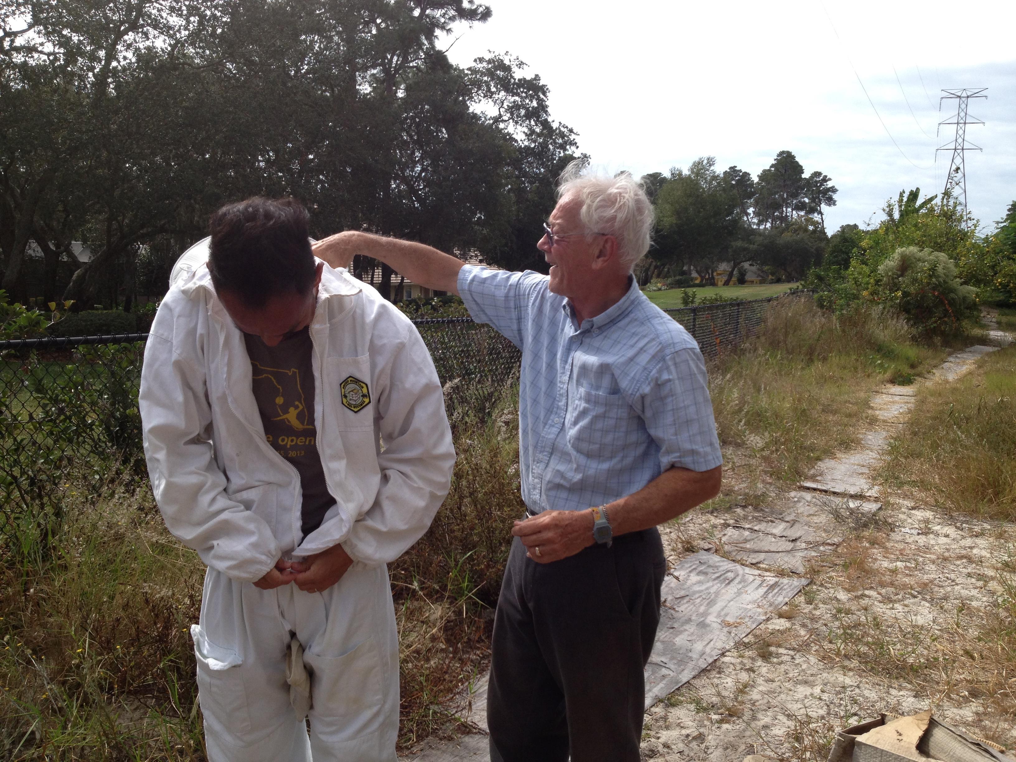 An elderly man instructs a young man in beekeeping techniques near a field on a sunny day.