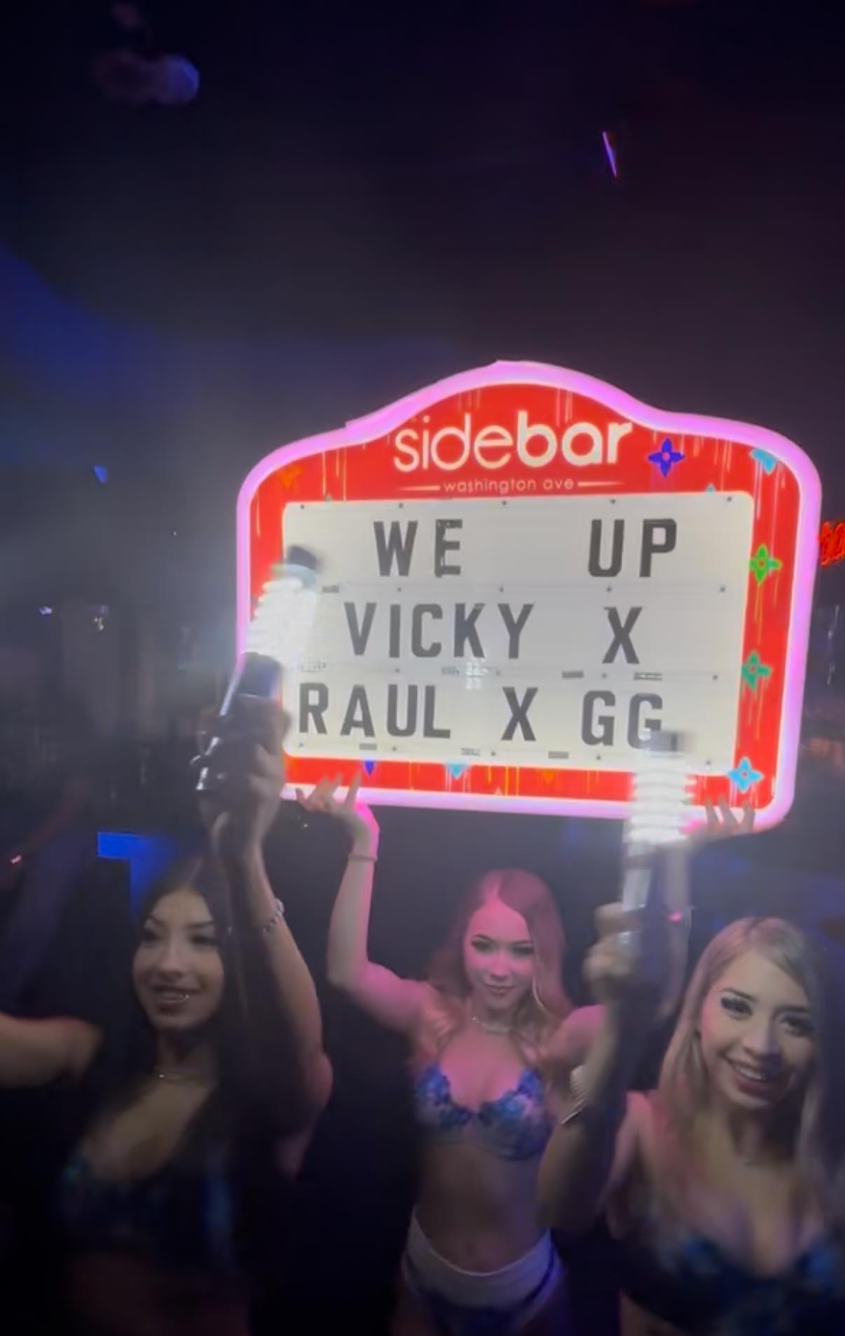 Group of friends celebrate at a vibrant nightclub, holding a bright neon sign with names.