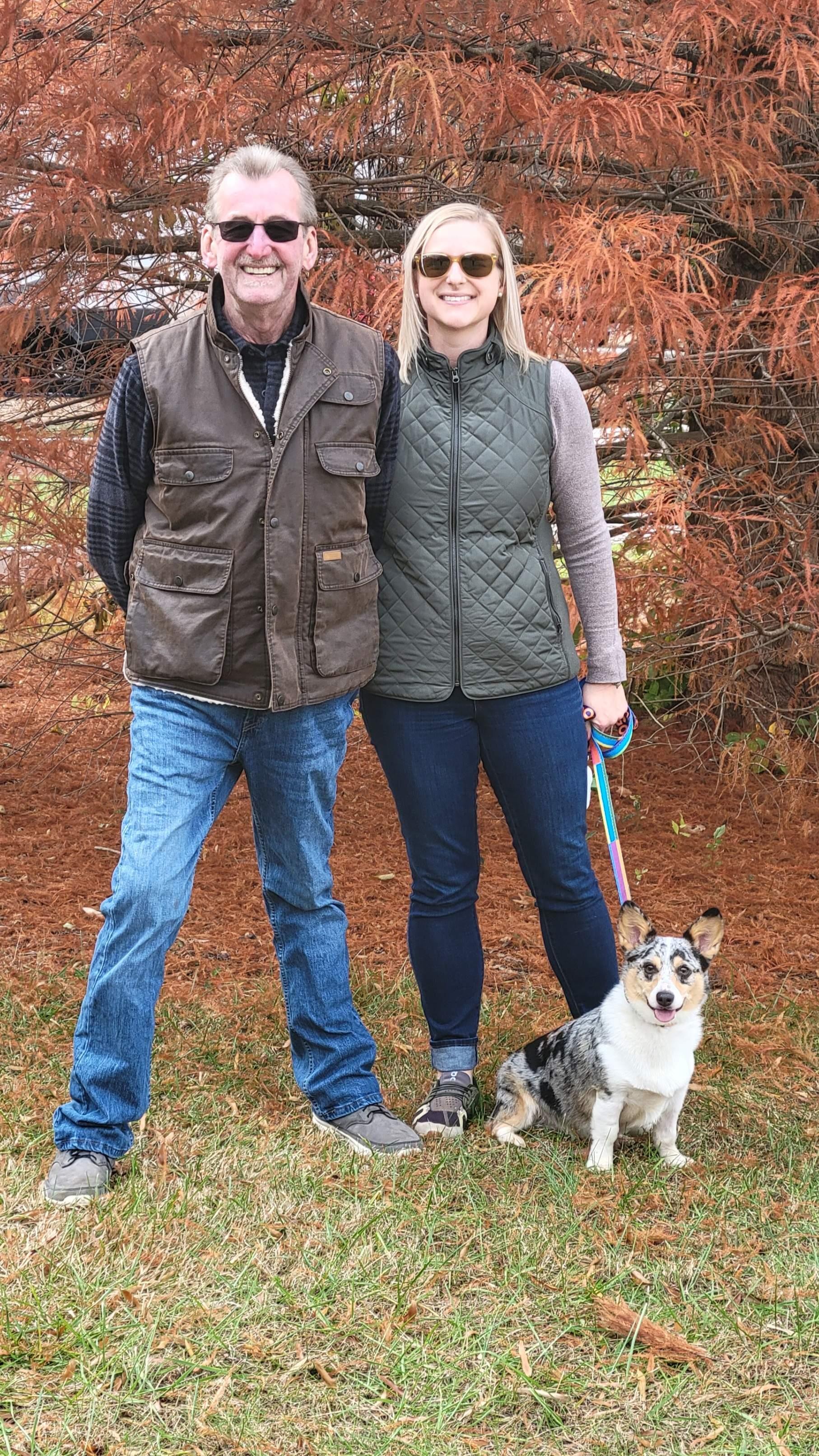 A couple stands together with their dog in a cozy outdoor setting surrounded by fall foliage.