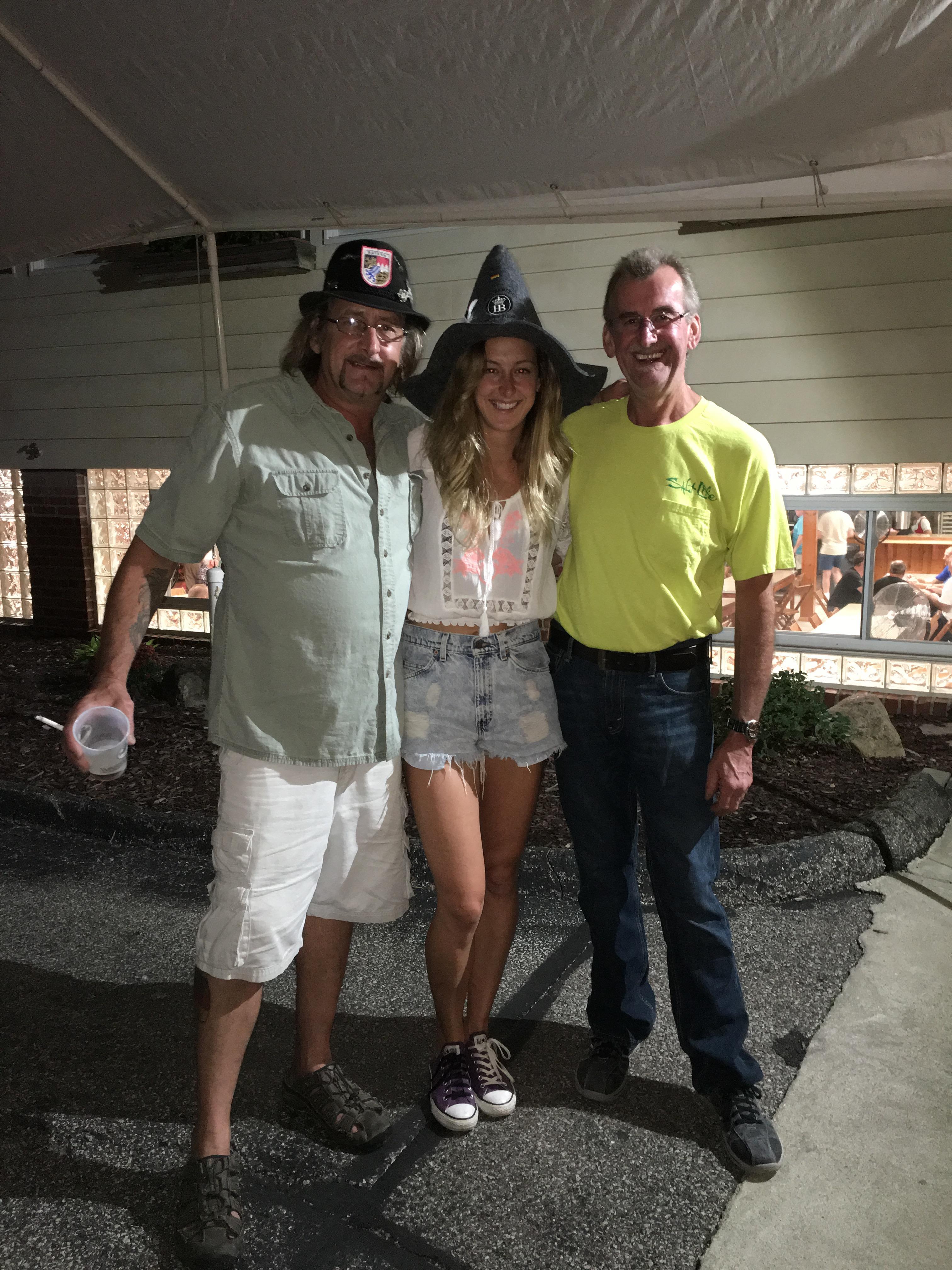 Three friends pose together, smiling, in casual outfits under outdoor lighting during summer.