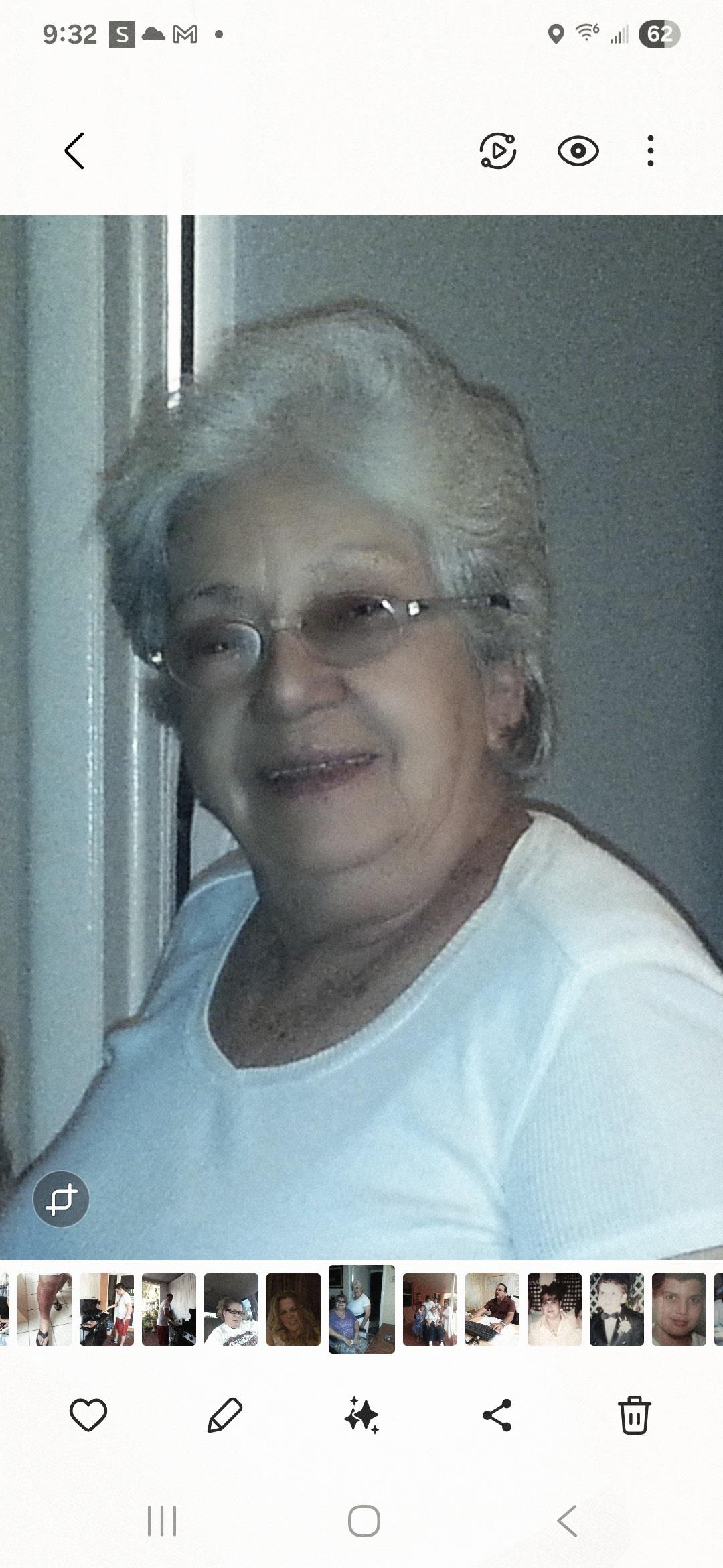 A cheerful elderly woman with glasses stands in a well-lit indoor area, exuding warmth and kindness.