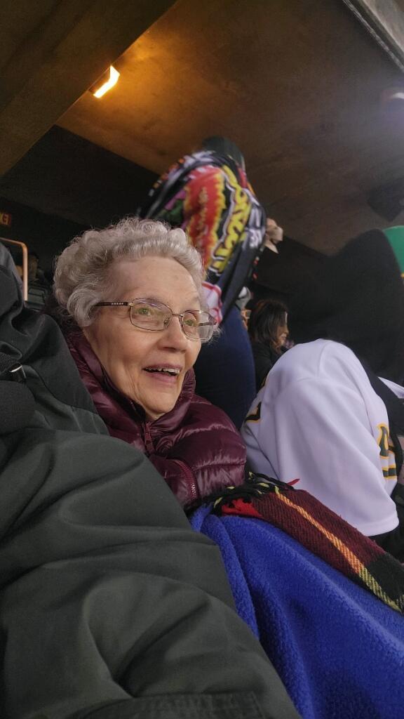 An elderly woman smiles joyfully while surrounded by lively fans at a stadium.