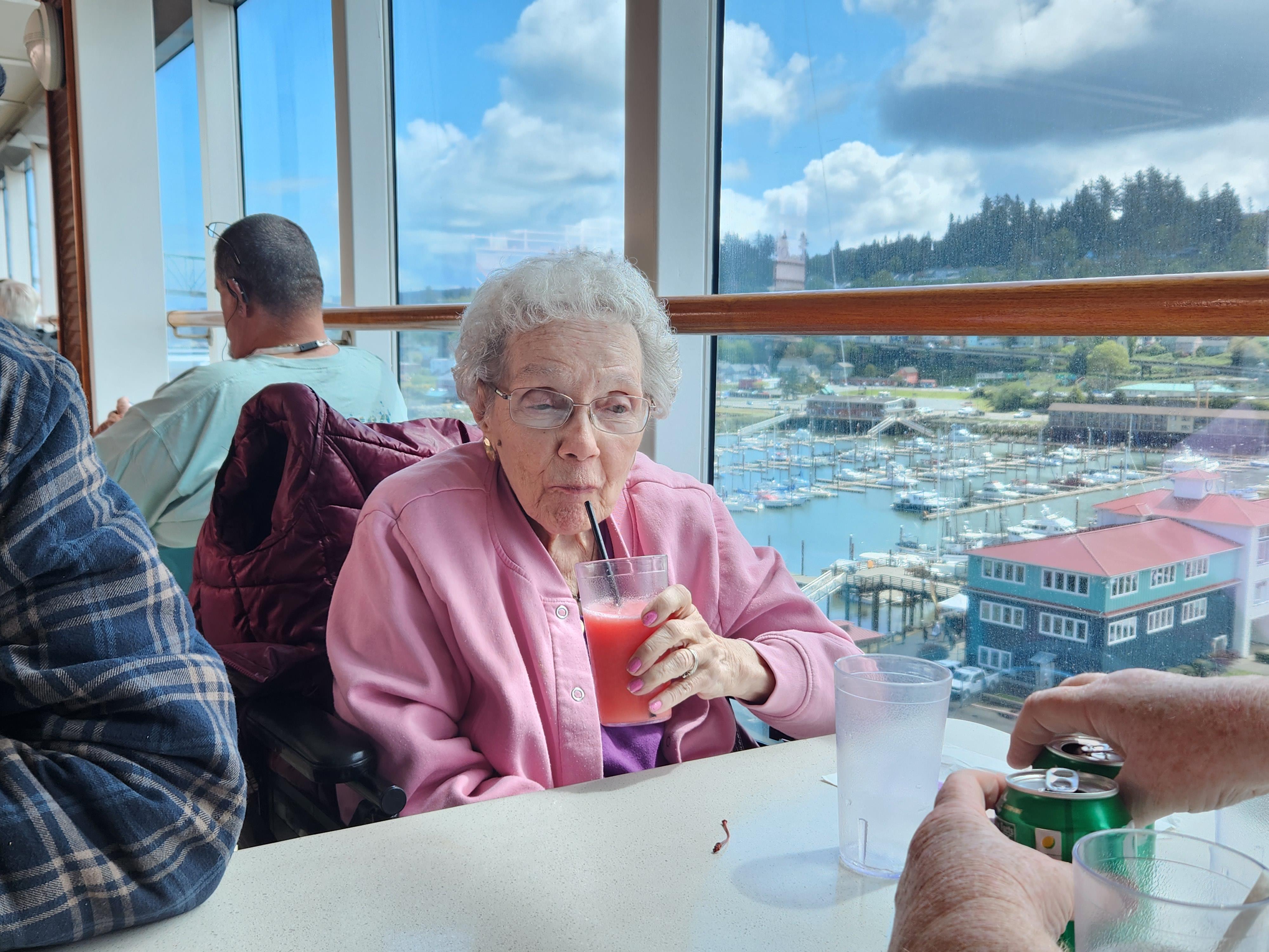 Senior woman drinks a fruity beverage while seated near large windows overlooking water.
