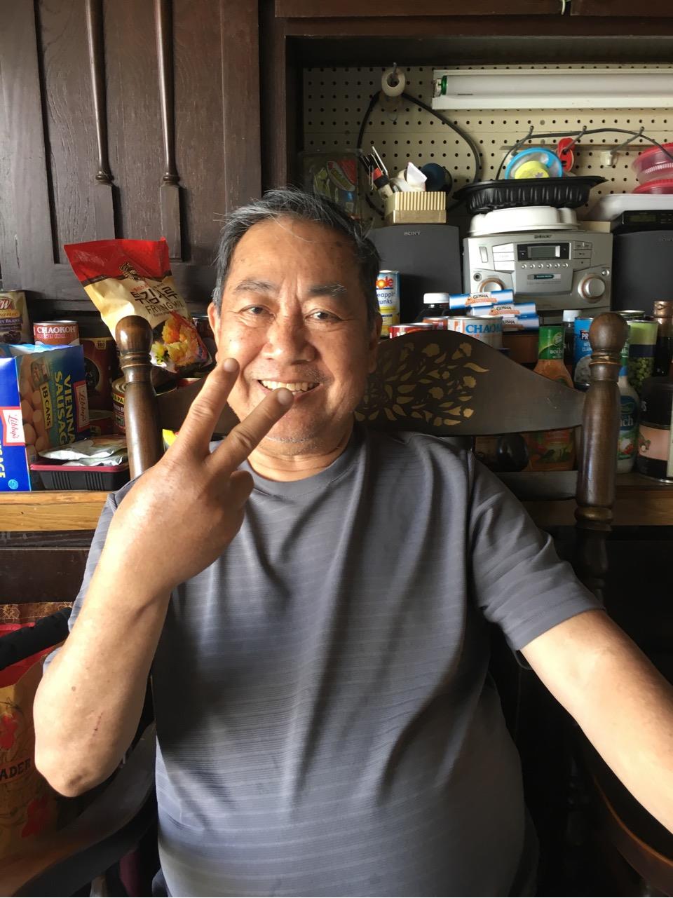 Smiling elderly man sits in a wooden chair, making a peace sign in a lively kitchen.