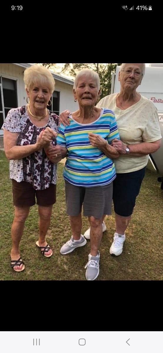 Three elderly women enjoy each other's company while standing outside in the warm sun.