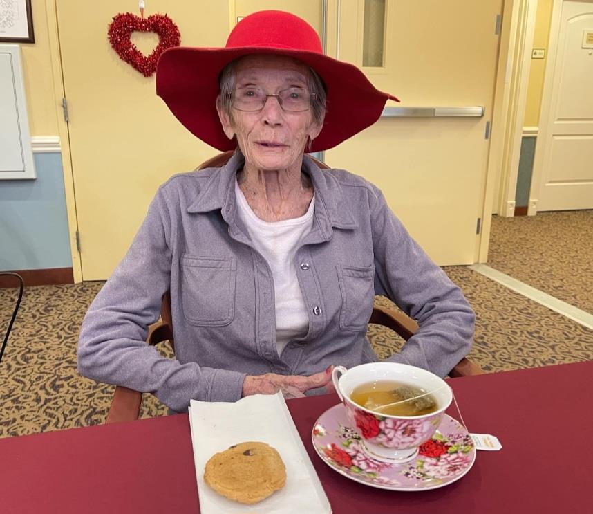 A senior woman sits at a table with a cup of tea and a cookie while smiling in a festive room.