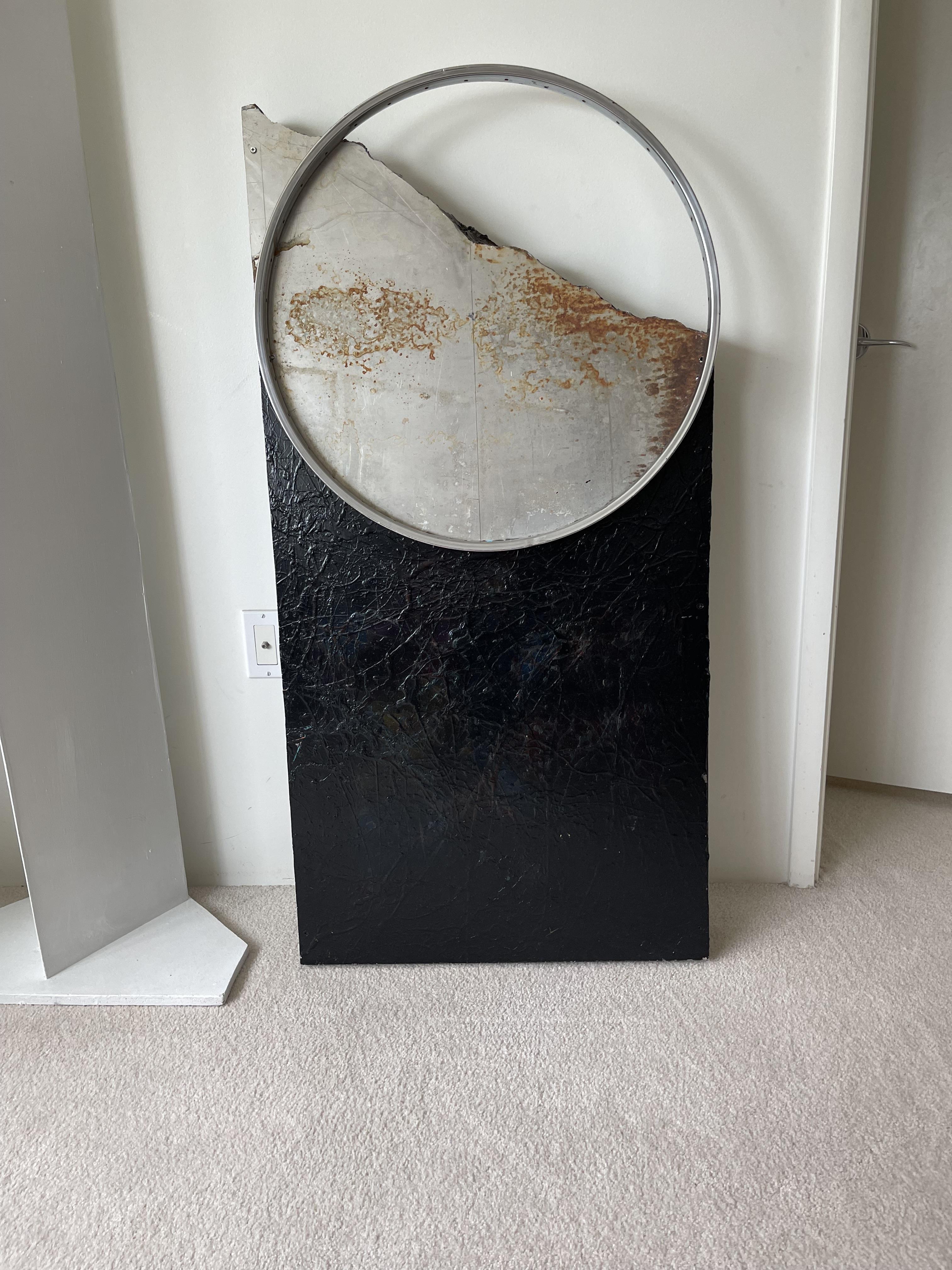 A black panel stands next to a wall. A round mirror rests above the panel in a quiet room.