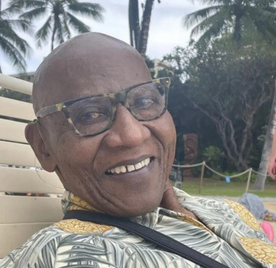 Man with glasses smiles while sitting in a chair surrounded by greenery and palm trees.