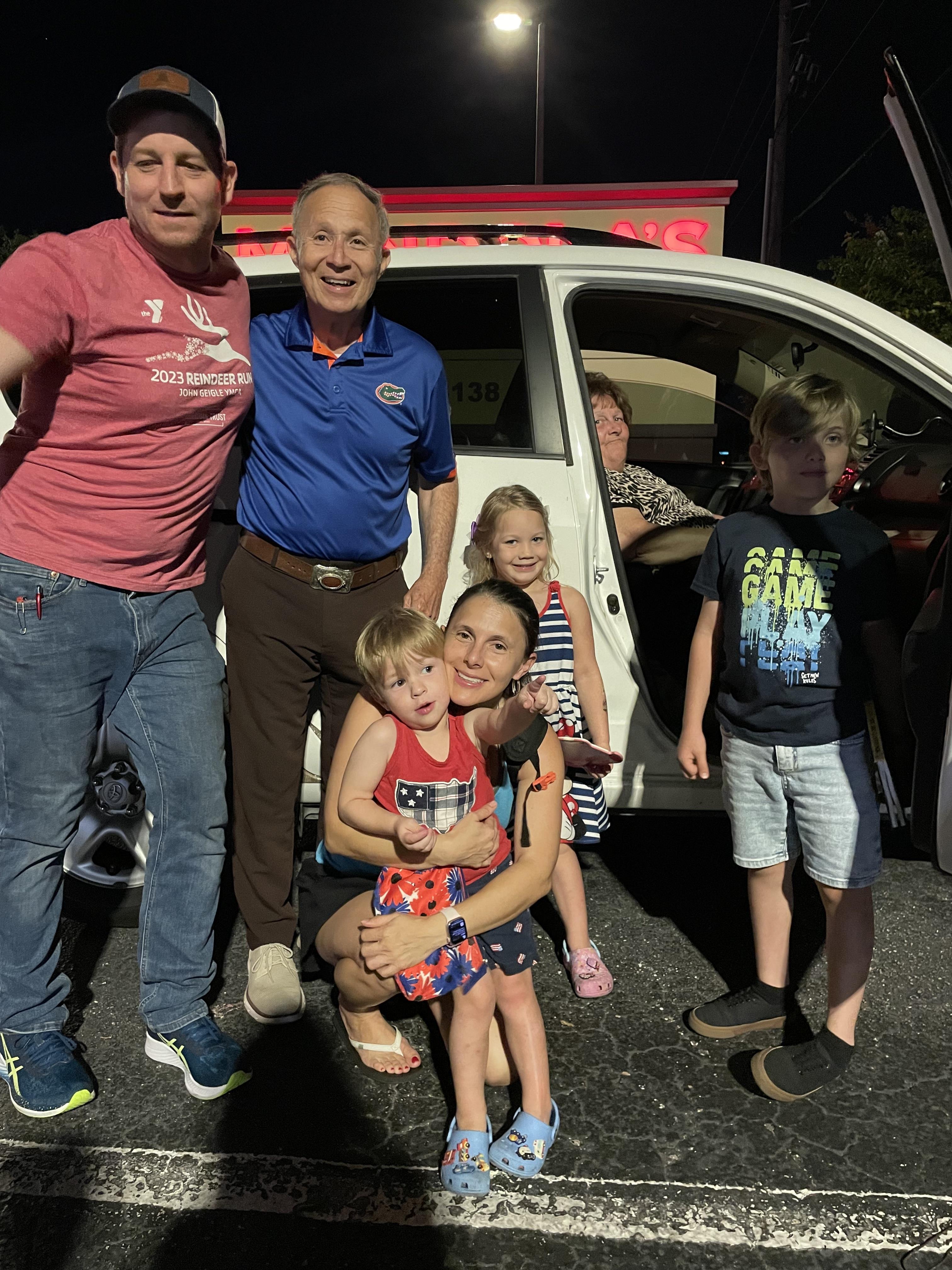 A lively group of adults and children enjoy a joyful moment outside a parked vehicle at night.